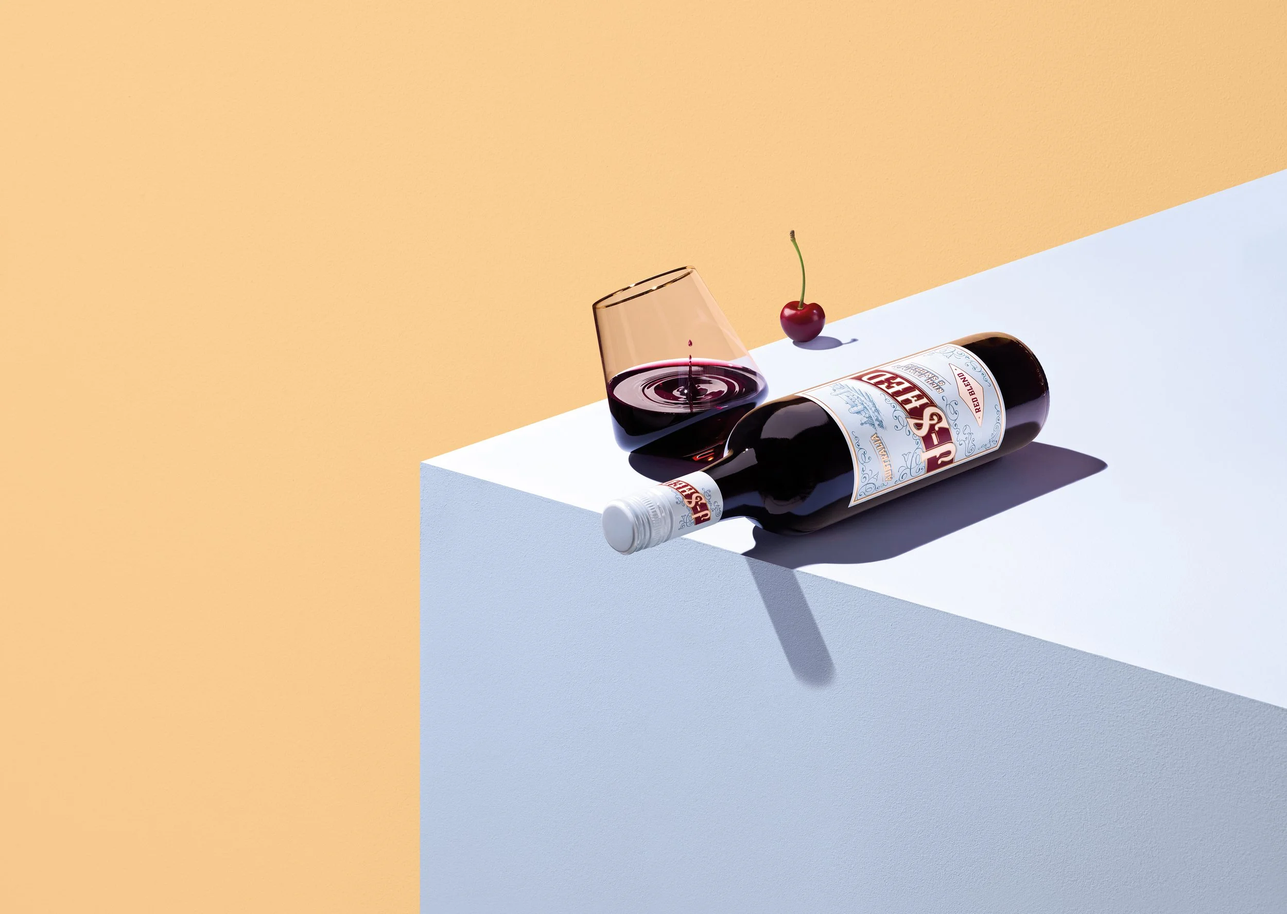 A bottle of red wine lying on its side, a wine glass filled with red wine, and a cherry with a green stem on a white surface against a two-tone background of beige and white.