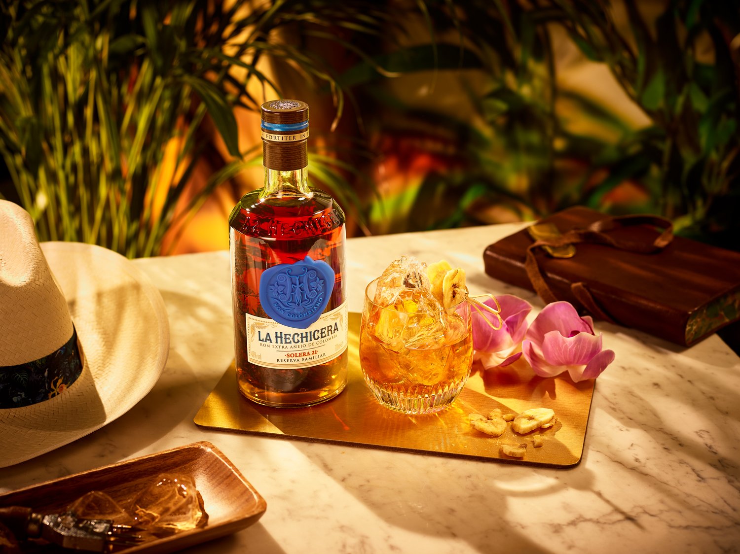 Bottle of La Hechicera rum, glass with ice and a yellow drink, pink flower petals, nuts, a straw, a hat, and a wrapped gift on a marble table with greenery in the background.