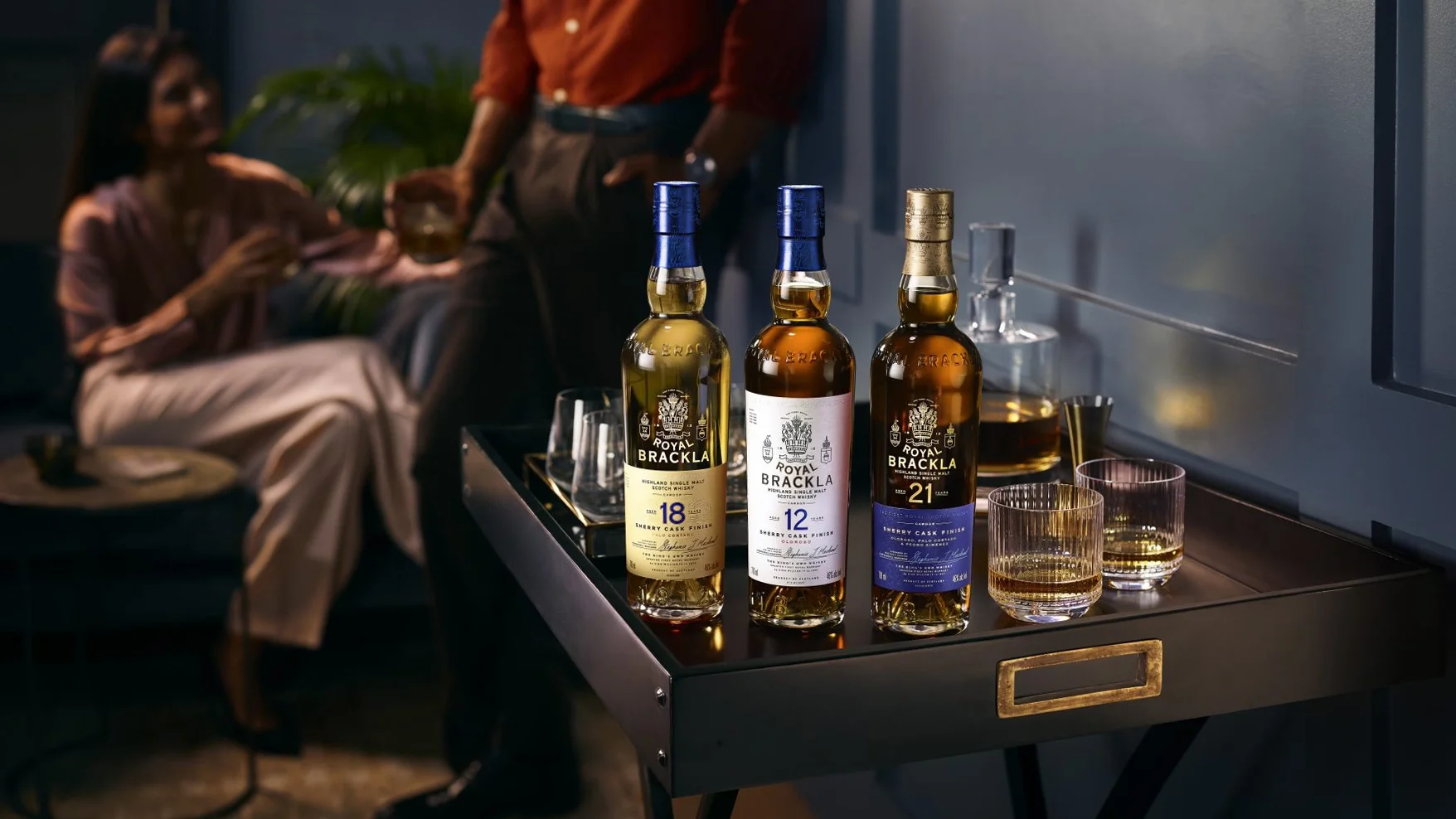 Three bottles of Royal Brackla Scotch whisky on a black bar cart, with glasses of whisky beside them. In the background, two people are sitting with drinks, engaging in conversation in a dimly lit room.
