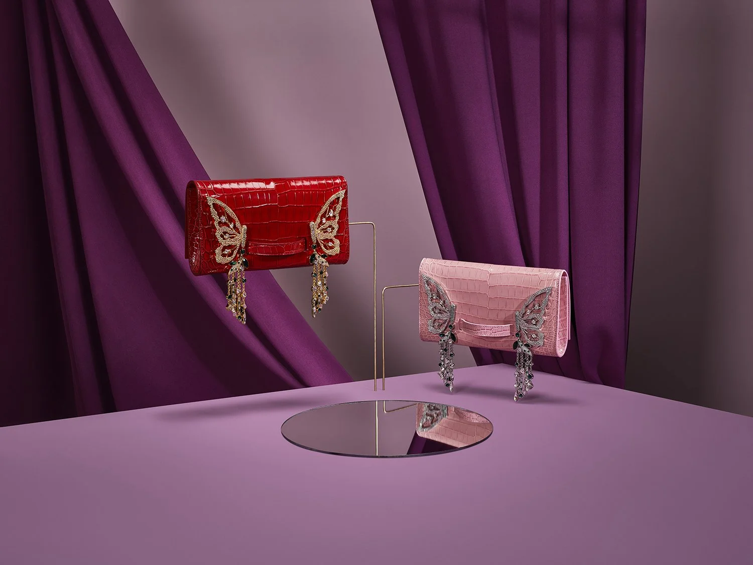 Two handbags with butterfly designs and dangling jewel embellishments are displayed on a purple backdrop, with a purple table and a round mirror reflecting part of the bags.