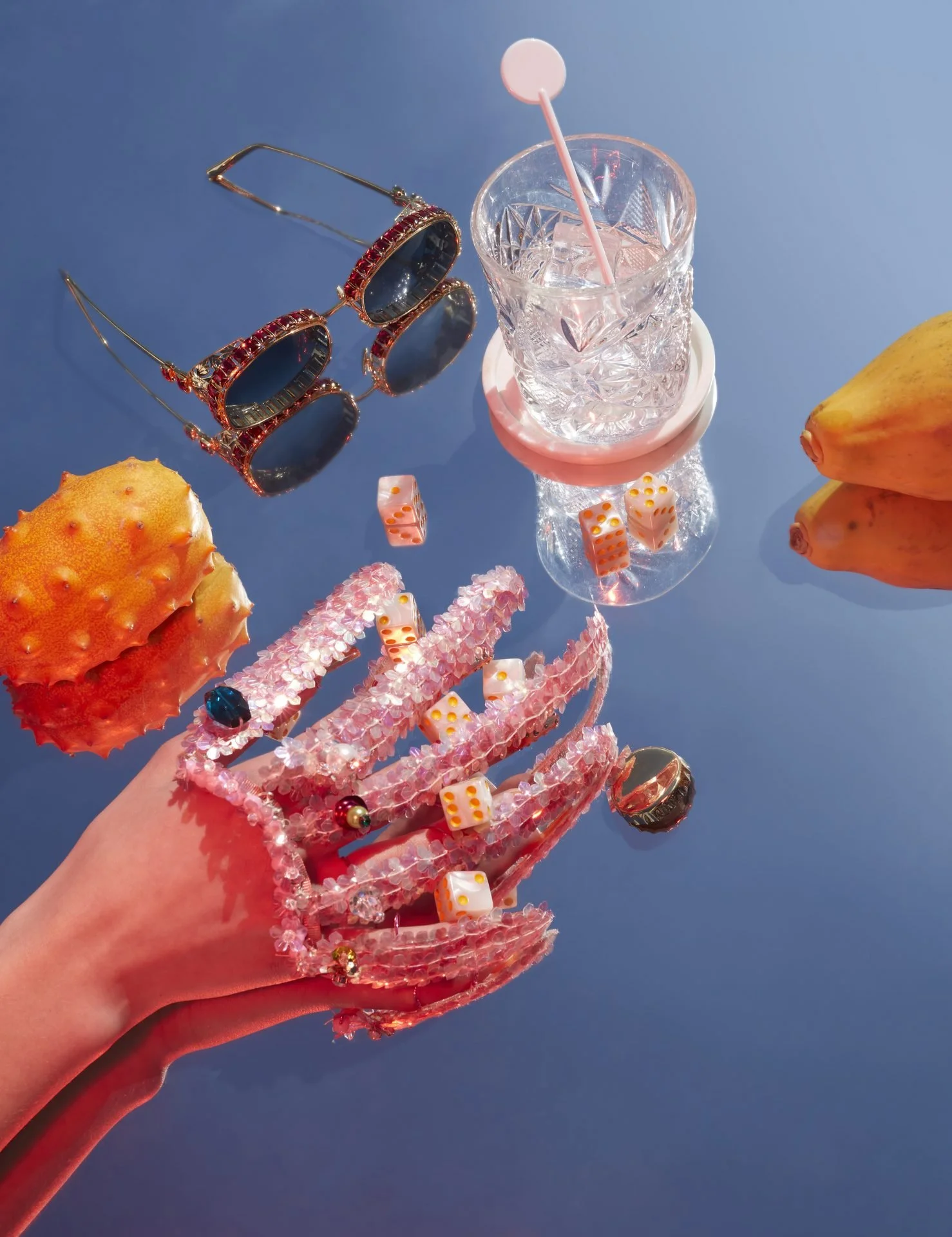 Colorful jewelry, sunglasses, and a glass of water with a straw are arranged on a blue surface with some fruits and dice nearby.