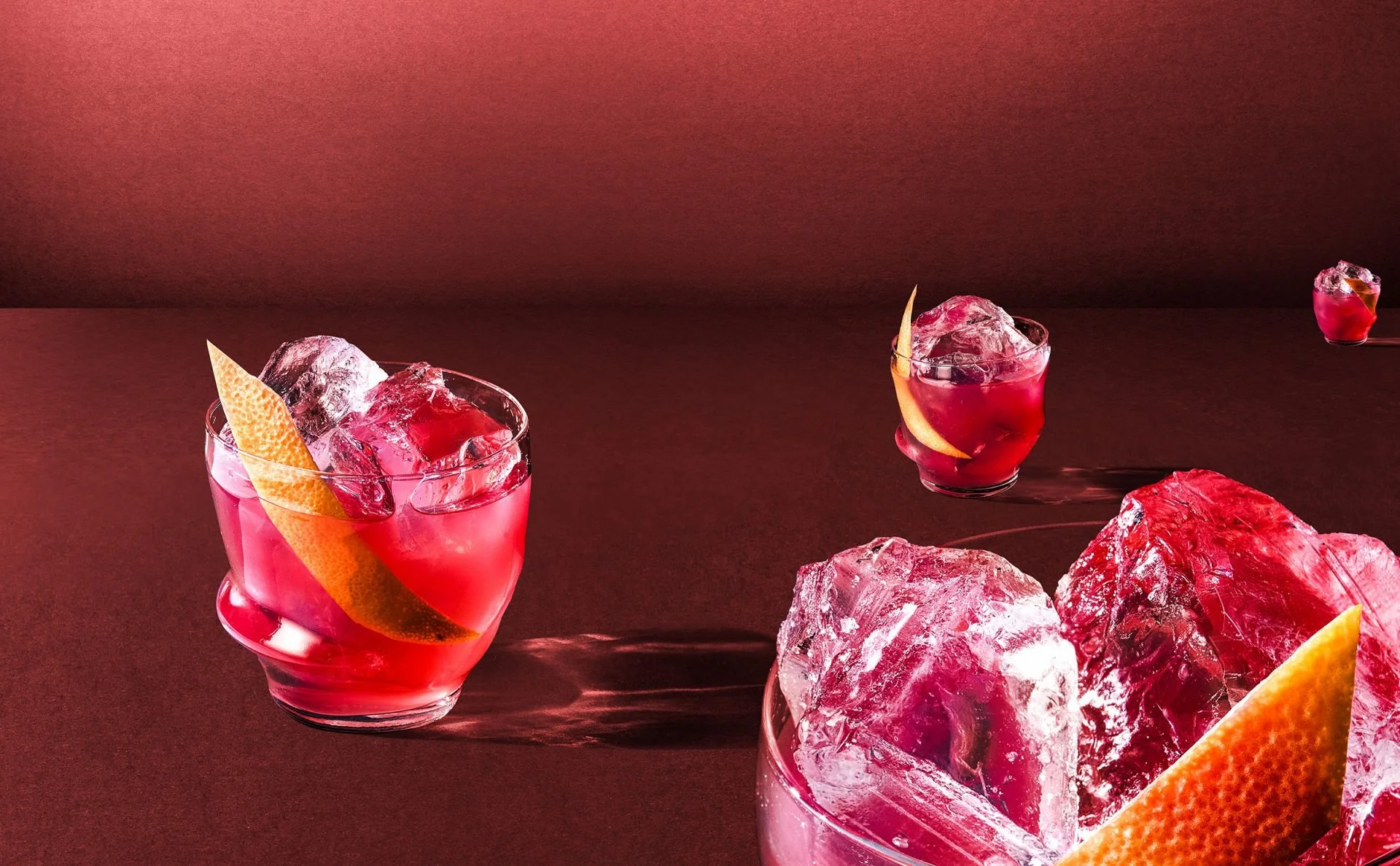 Three glasses of pink cocktail with ice and orange slices on a dark surface, with large ice rocks nearby, and a dark pink background.