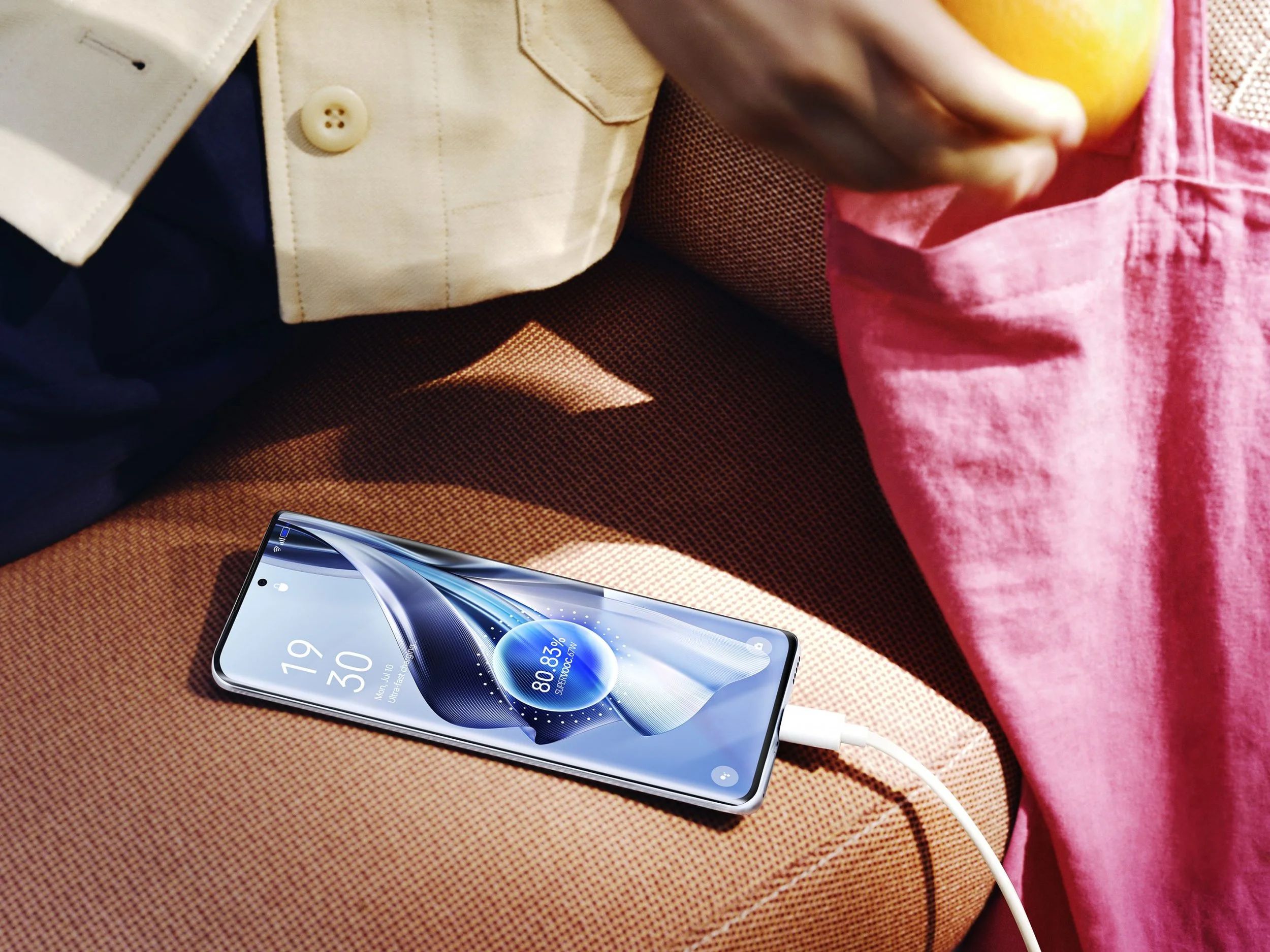 Close-up of a smartphone with a digital display showing the time as 19:30 and battery at 80%, connected to a charging cable. The phone is resting on a textured brown surface, next to a person wearing pink pants and a yellow shirt, with part of a person's cream-colored jacket visible in the background.