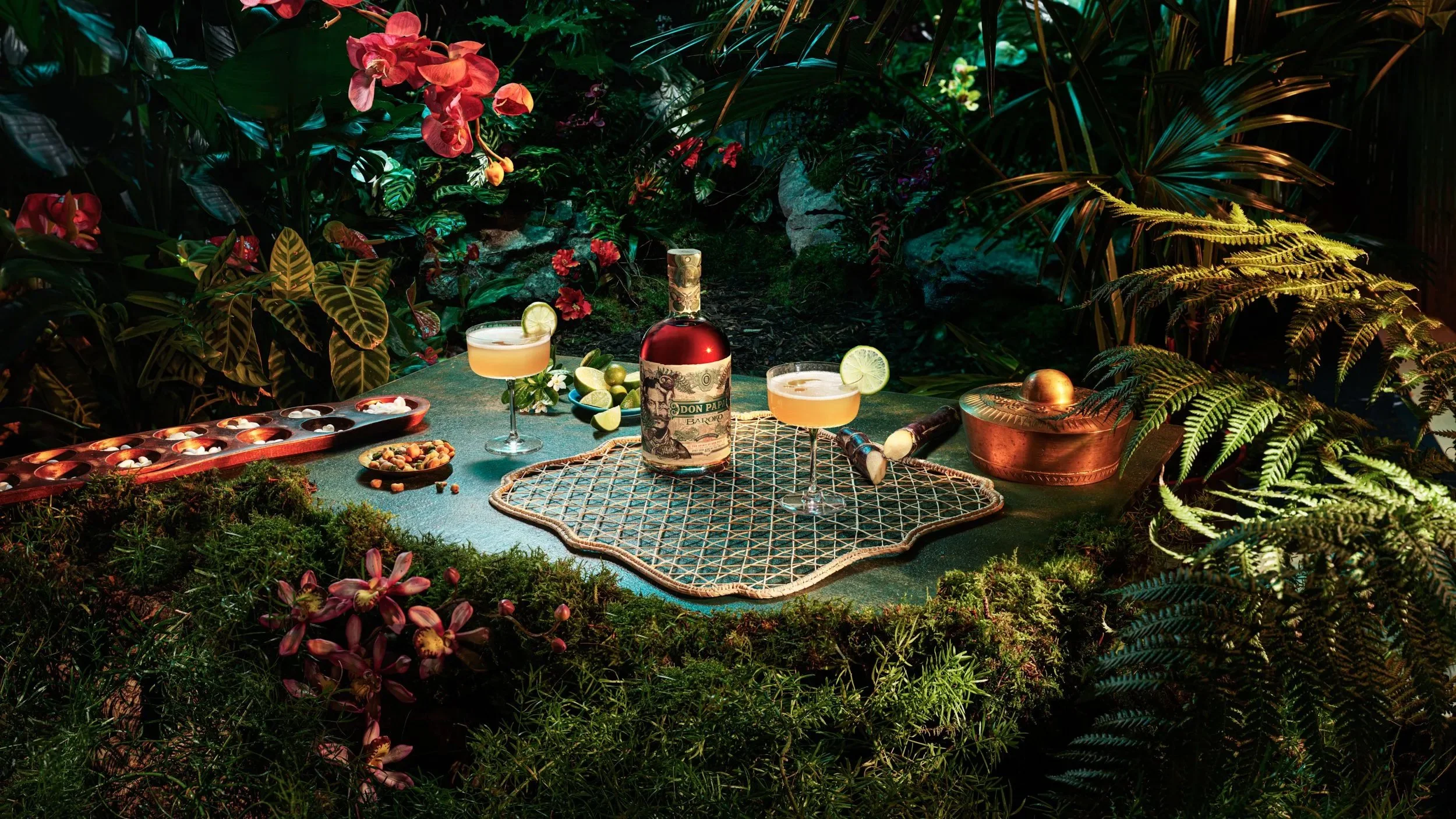 Tropical outdoor setting with a green table, two cocktails garnished with lime slices, a bottle of Don Papa rum, a tray of chocolates, and scattered nuts, surrounded by lush tropical plants and flowers.