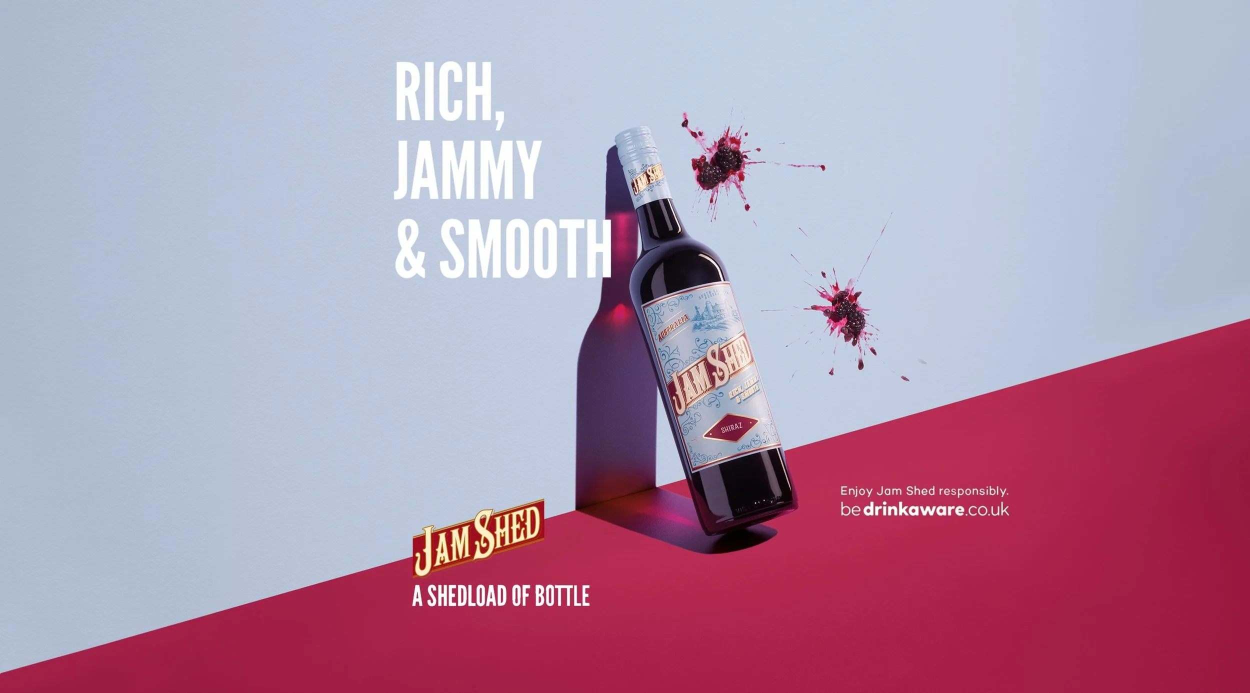 A bottle of Jam Shed Shiraz wine with a splash of blackberries and red wine splattered behind it, against a light blue and red background, with text promoting responsible drinking.