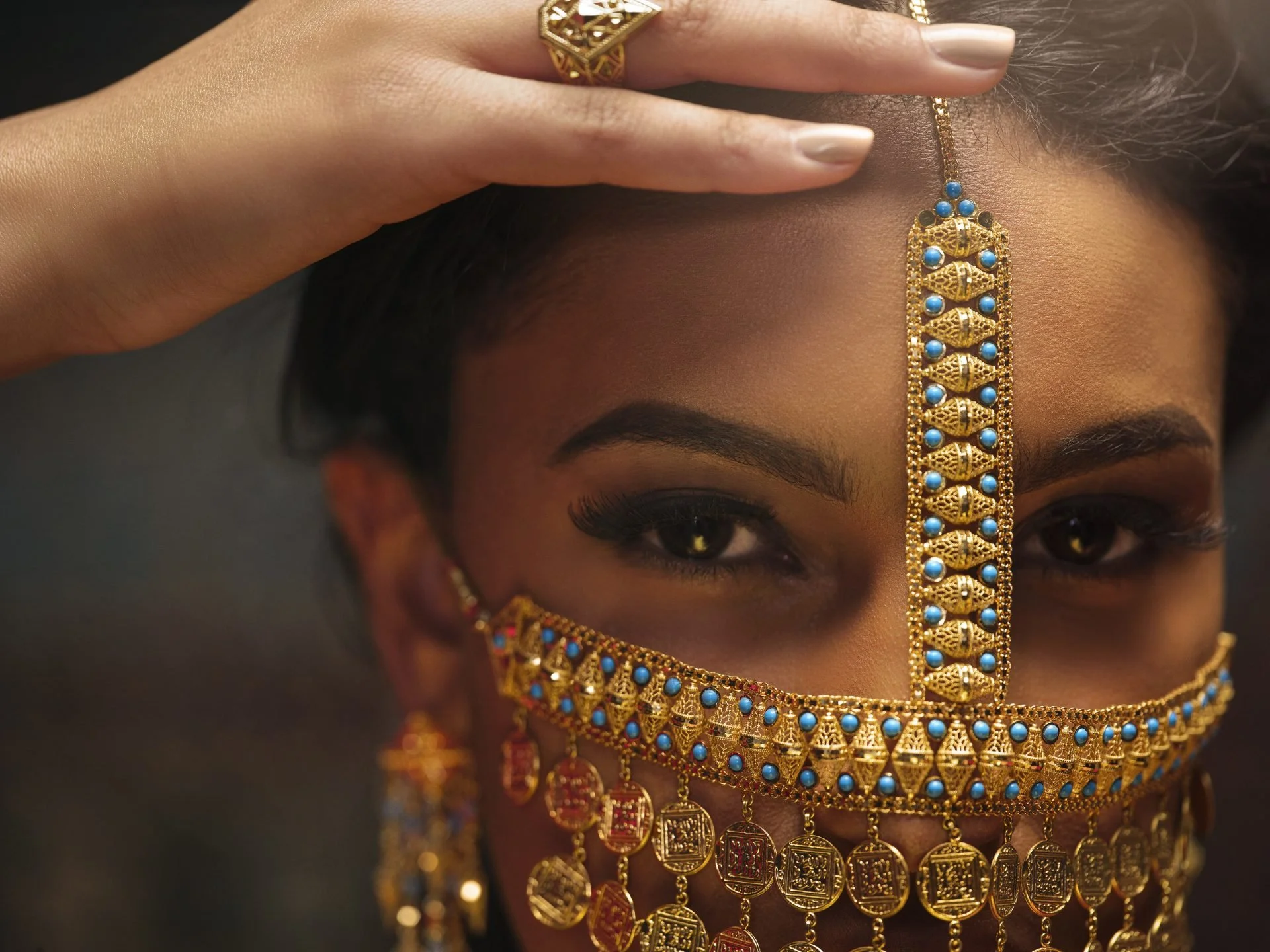 Close-up of a woman's face adorned with intricate gold and turquoise jewelry, including a forehead chain, earrings, and necklaces.