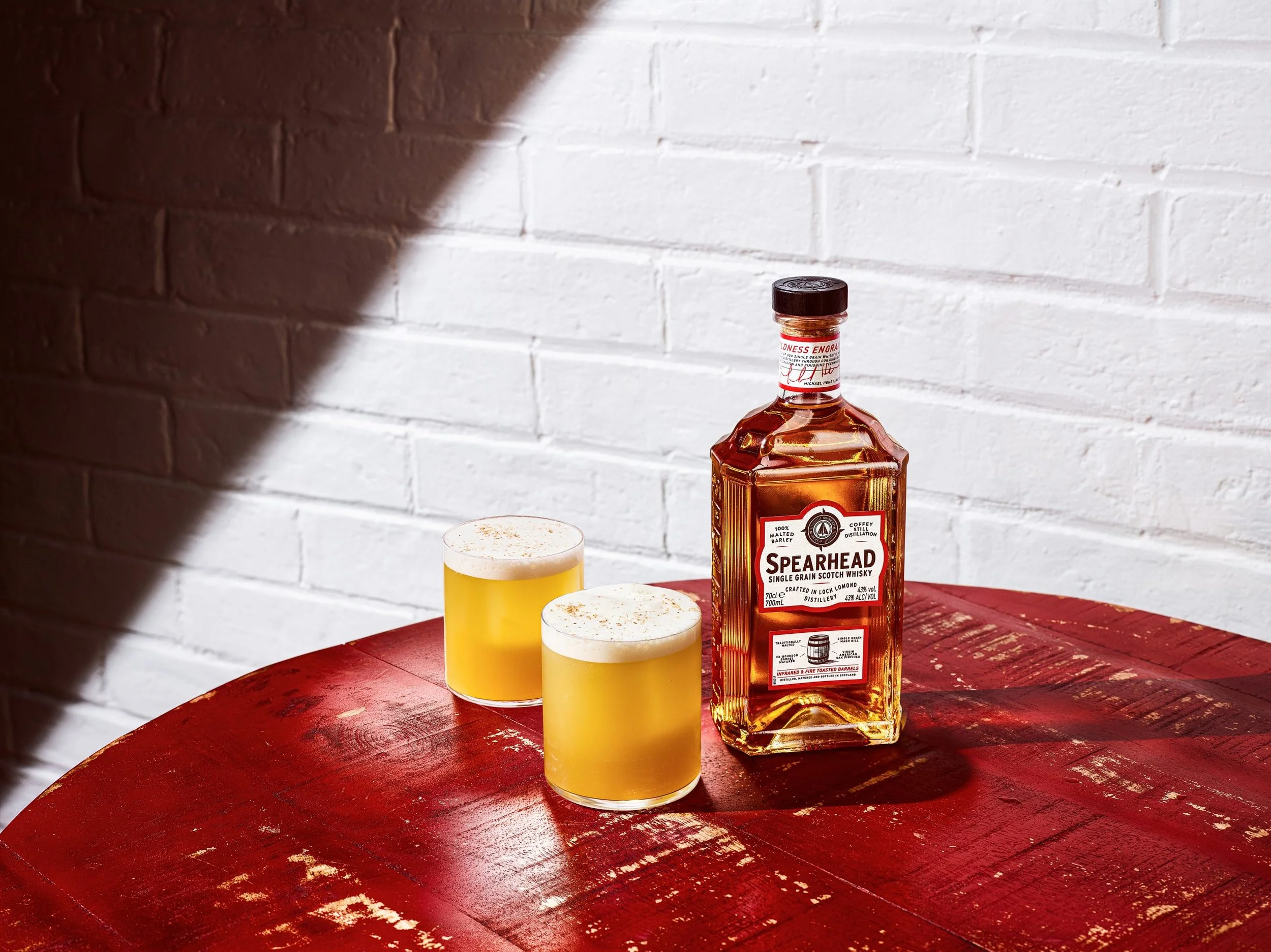 A bottle of Spearhead single grain Scotch whisky on a red wooden table, with two drinks topped with foam in yellow glasses, against a white brick wall background.