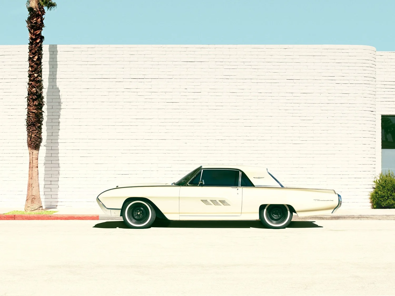 A vintage cream-colored car parked on a sunny street in front of a white brick wall. There is a palm tree on the left and a small bush on the right.