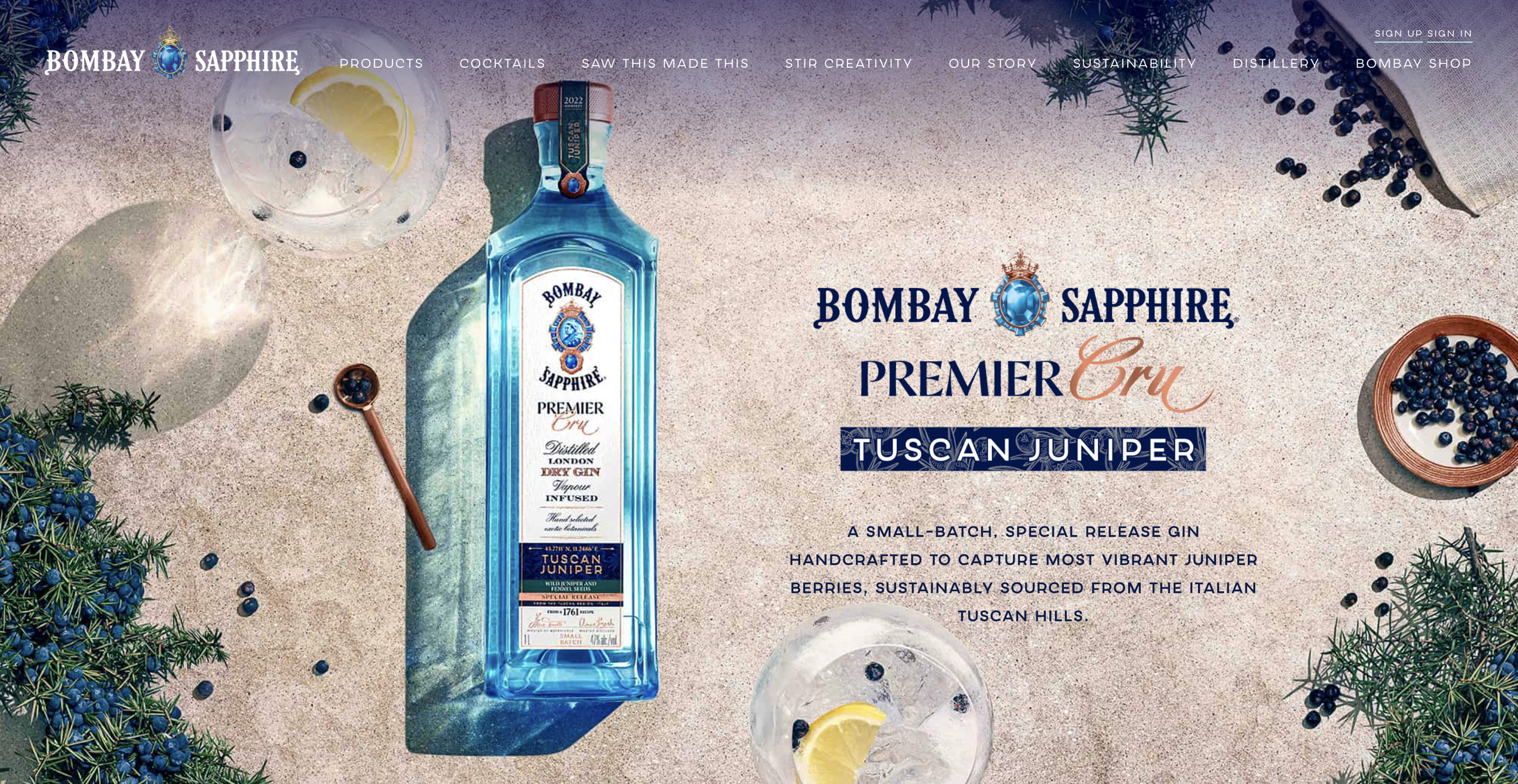 Advertisement for Bombay Sapphire Premier Cru gin, featuring a blue gin bottle on a sandy surface surrounded by juniper berries, lemon slices, and ice glasses, with text about the product's features and sourcing.