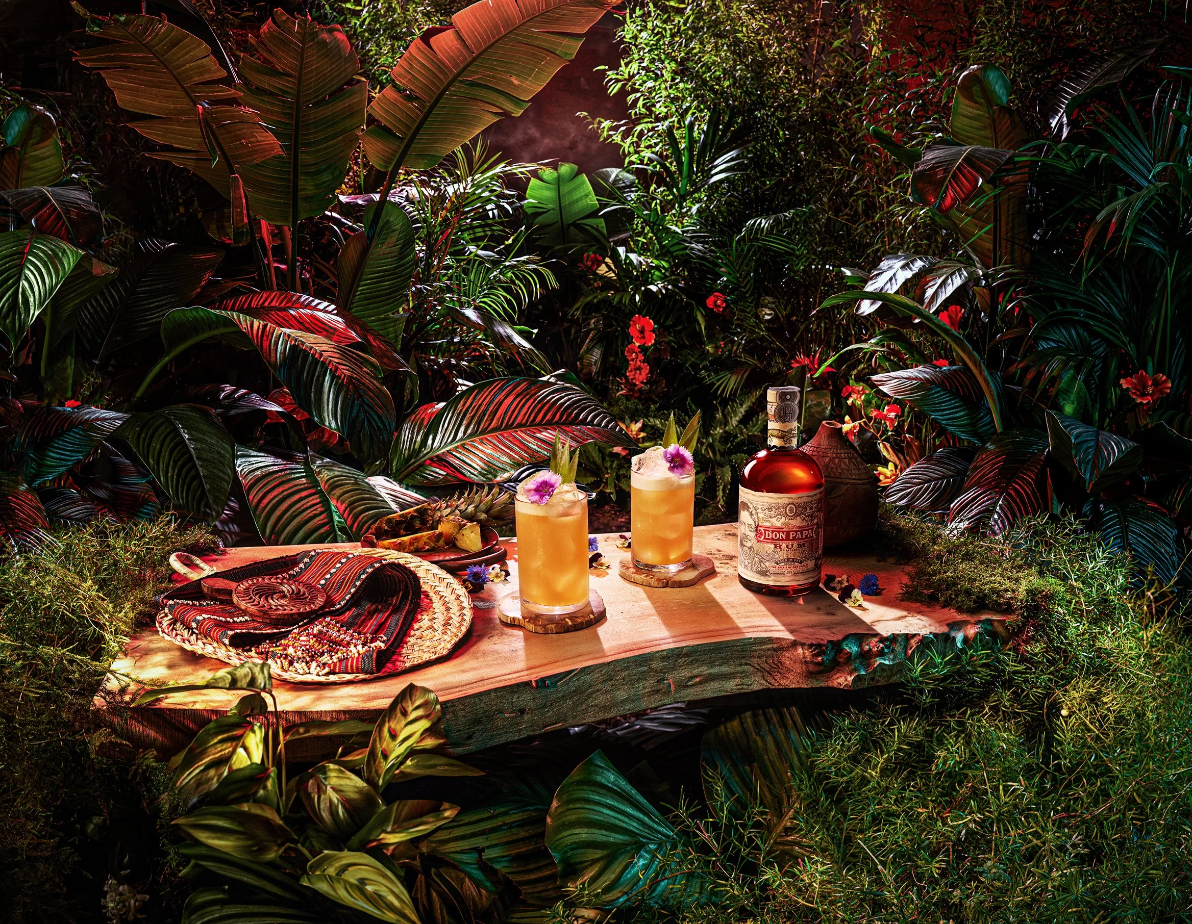 Tropical jungle scene with large green and red leaves, red flowers, and a table set with drinks, snacks, and a bottle of Don Papa rum.