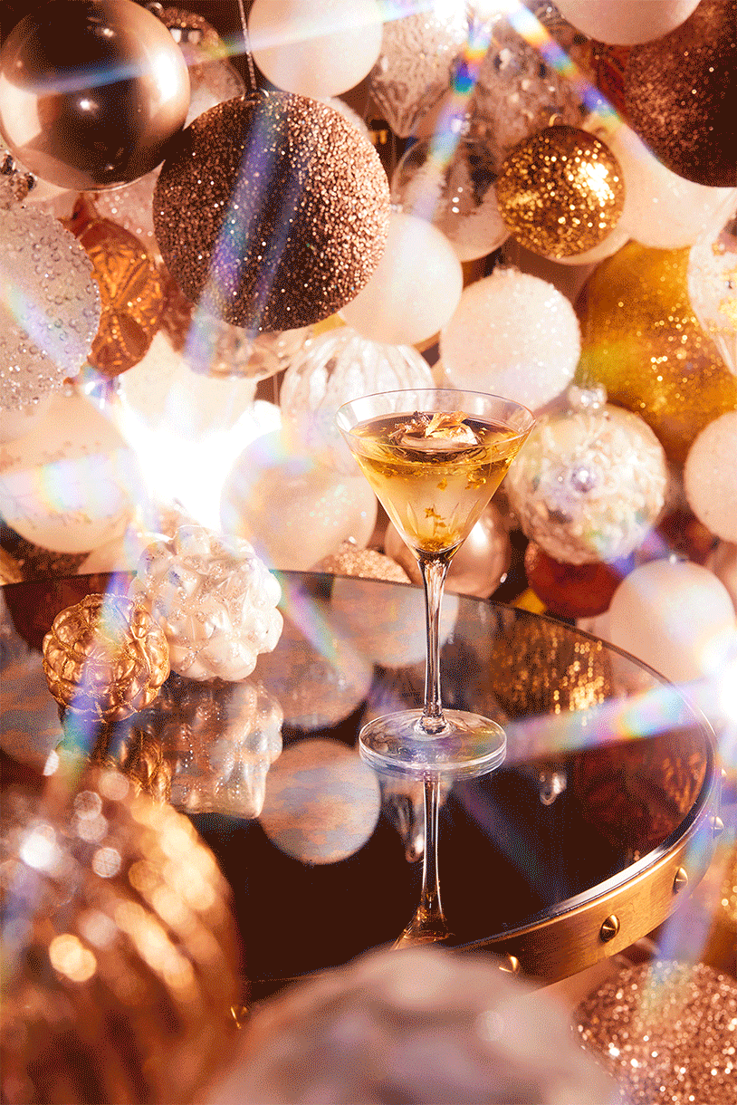 A glass of champagne on a reflective surface surrounded by white, gold, silver, and glittery ornaments and holiday decorations with bright, colorful light reflections.