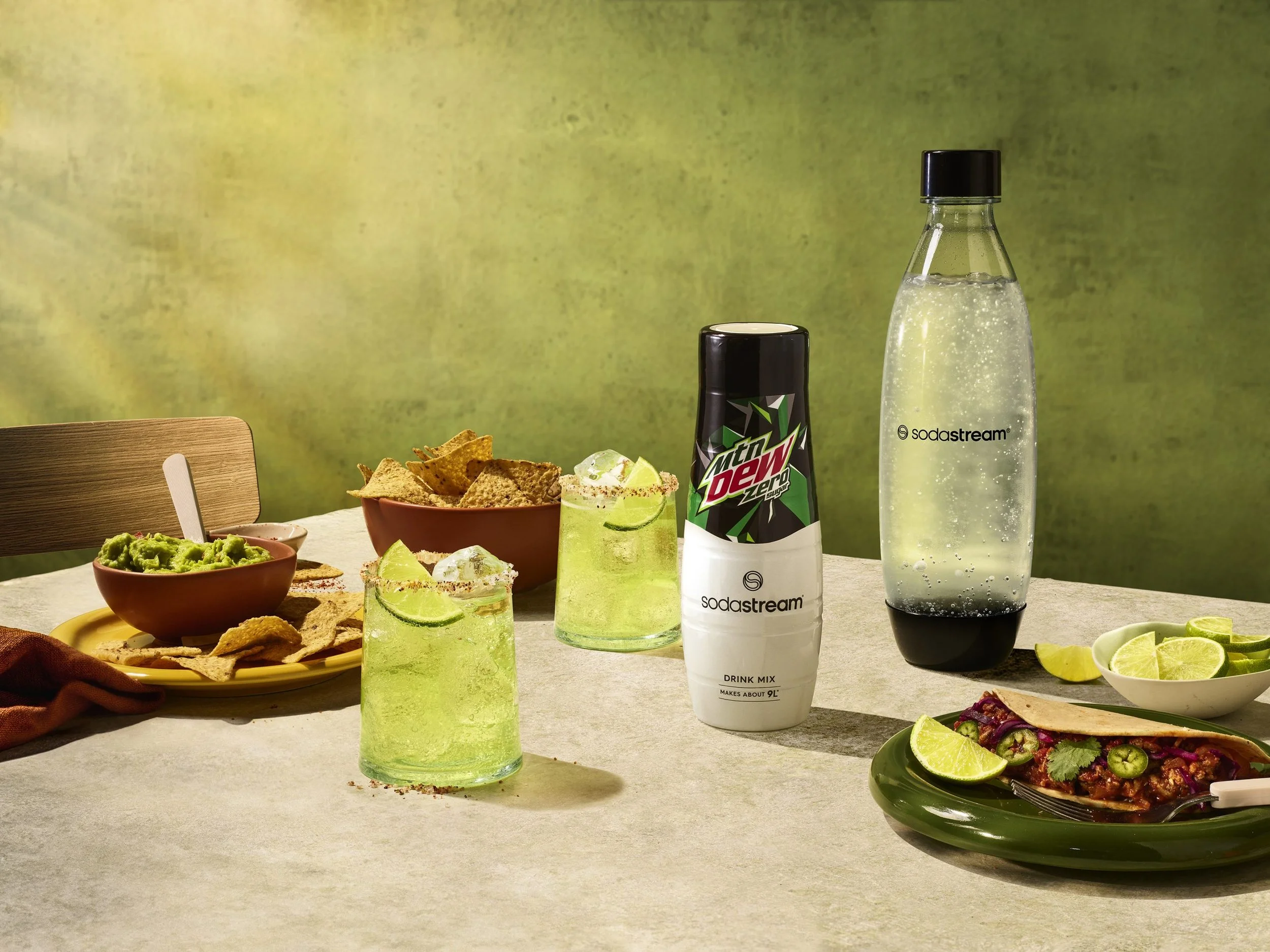 A table with snacks and drinks including lime wedges, guacamole, chips, tacos, seltzer water, and Mountain Dew Zero Sugar in a black can, set against a green textured background.