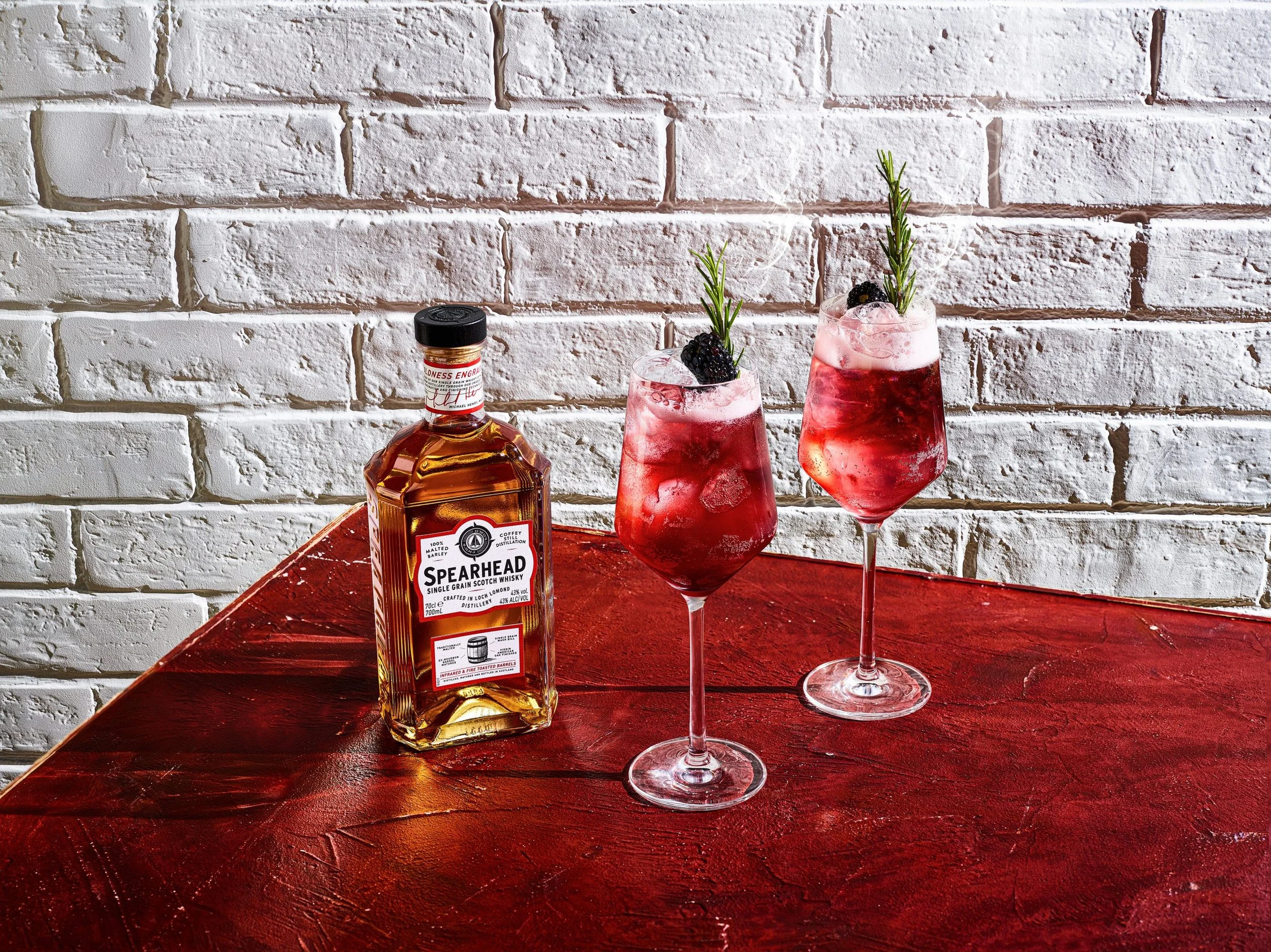 A bottle of Spearhead single grain Scotch whisky and two pink cocktails garnished with blackberries and sprigs of rosemary on a red wooden table against a white brick wall.