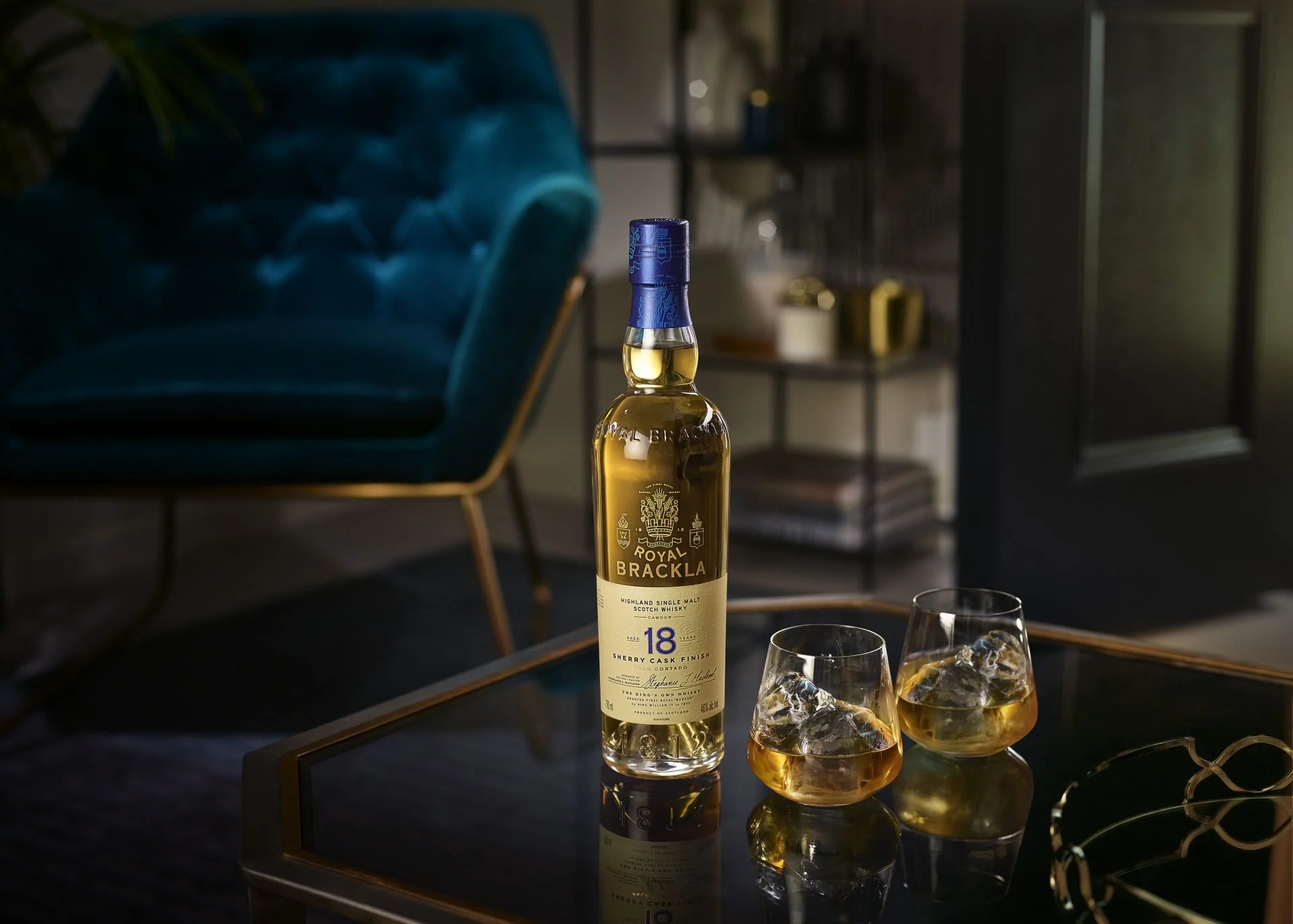 A bottle of Royal Brackla 18-year-old single malt Scotch whisky on a glass table with three glasses of whisky with ice cubes. The background features a teal velvet armchair and a blurred bookshelf in a modern living room.
