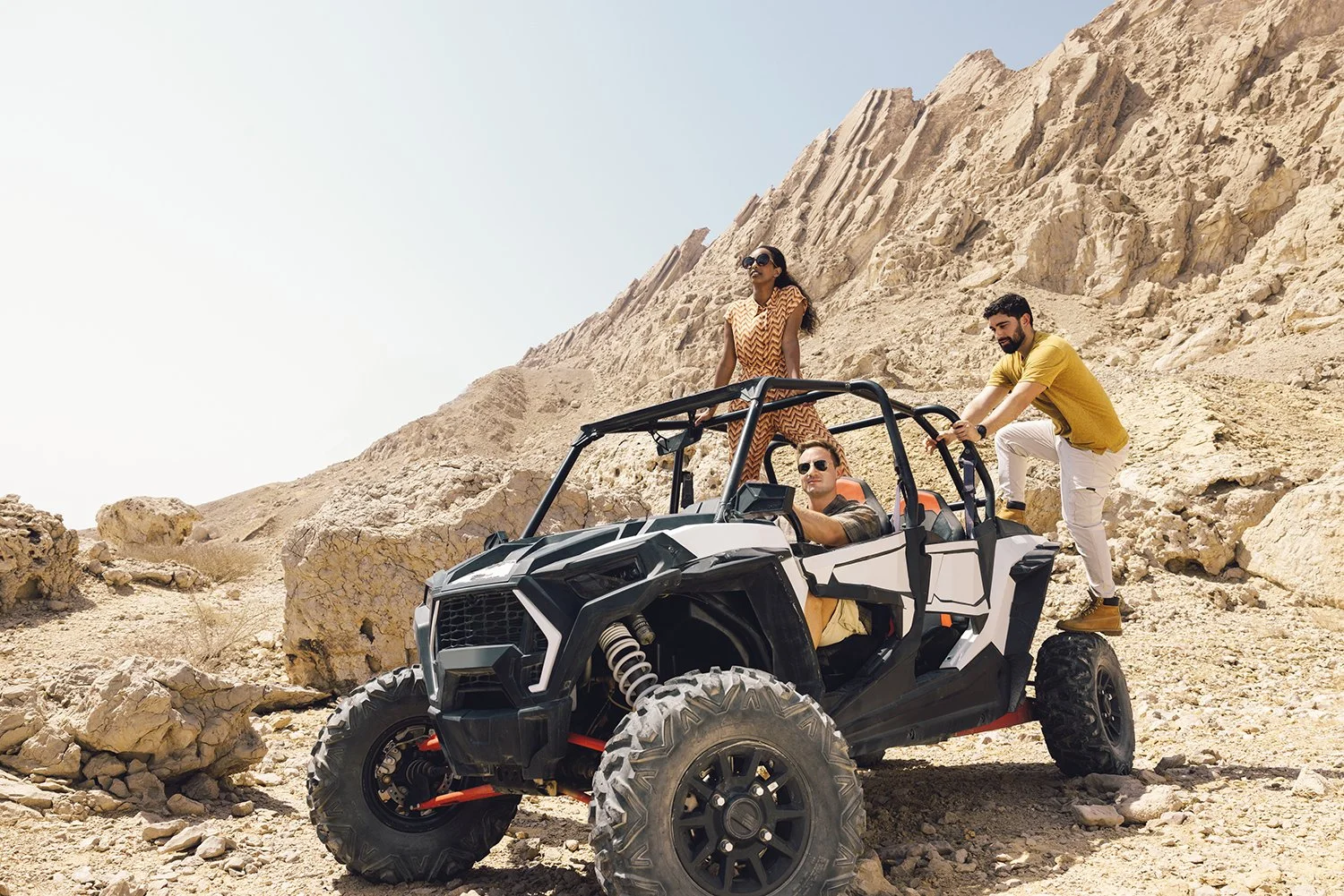 Three people in a desert mountainous area with a rugged off-road vehicle, one person is sitting inside the vehicle, and two others are standing on the vehicle's side, with rocky terrain in the background.