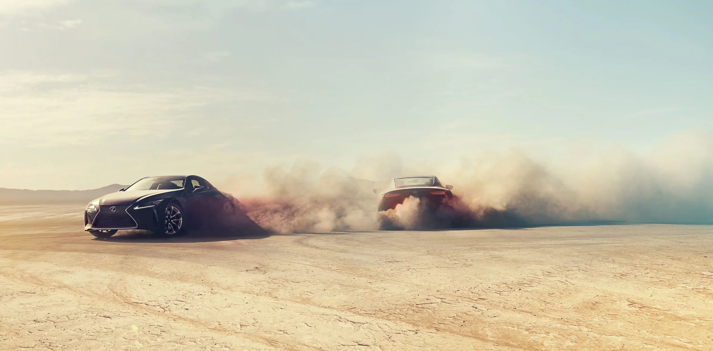 Two luxury sports cars, a black Lexus and a silver convertible, race across a dry, flat desert landscape with dust and smoke trailing behind them on a clear day with a blue sky.
