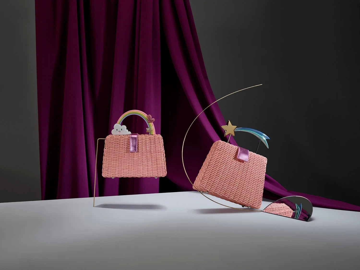 Pink woven handbags with decorative rainbow, cloud, star, and shooting star embellishments, displayed against a dark background with purple curtains.