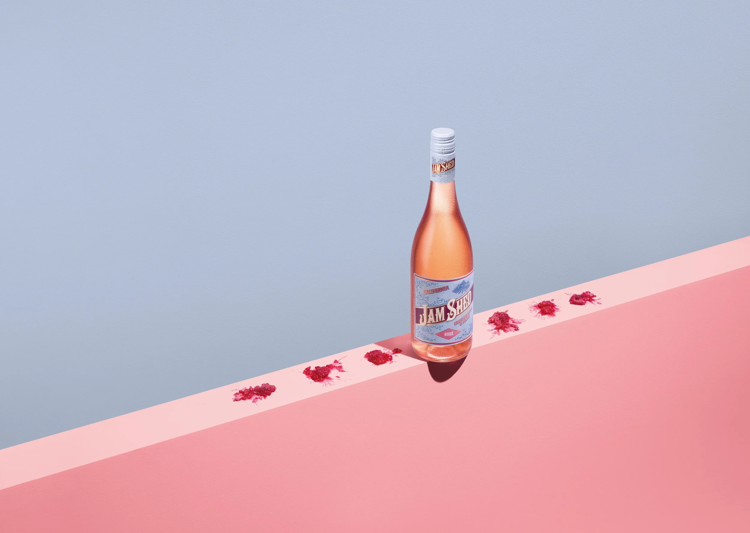 A pink bottle of rosé wine, labeled 'Jam Shed,' is placed on a pink surface with red raspberry-like objects scattered around. The background includes a blue wall.