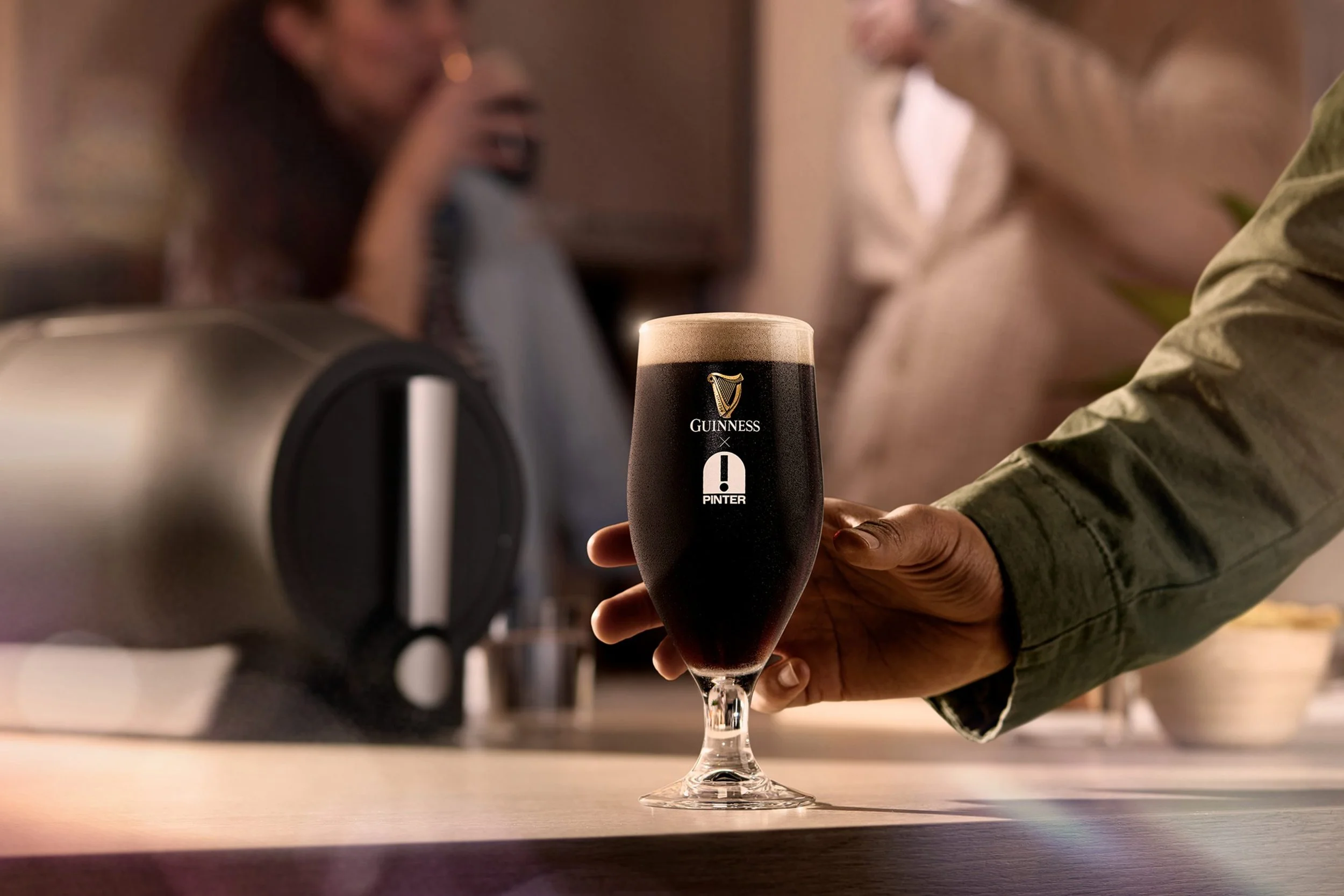 Close-up of a person holding a Guinness pint glass with dark beer and a foamy head, with the Guinness logo visible, in a bar or pub setting.