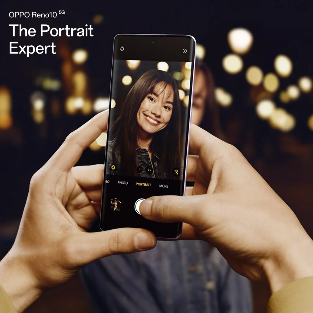 A person holding a smartphone taking a portrait photo of a smiling woman with long dark hair, in an indoor setting with blurred warm lights in the background.