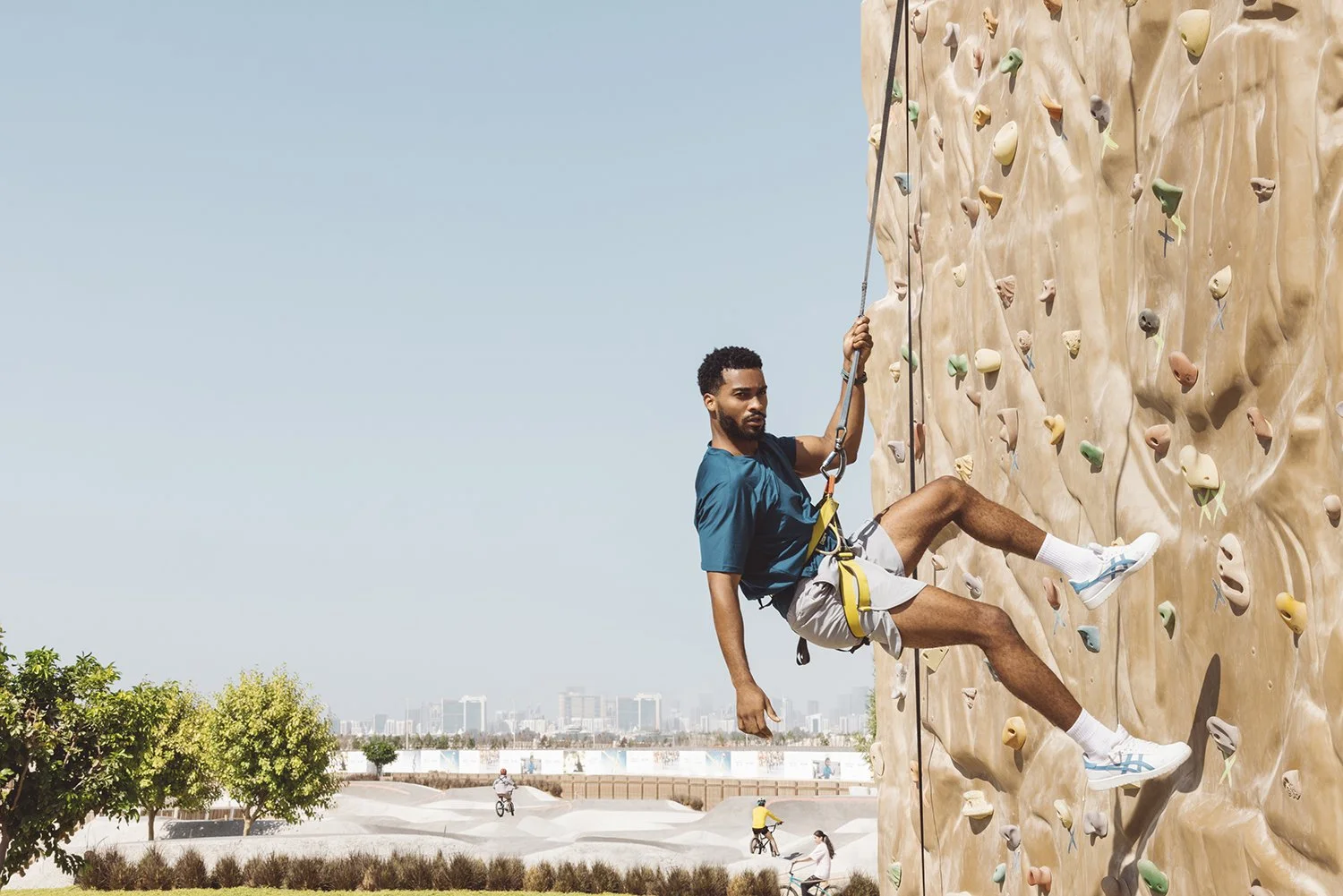 A man rock climbing on an outdoor artificial climbing wall, wearing a harness and athletic clothing, with cityscape and people riding bicycles in the background.
