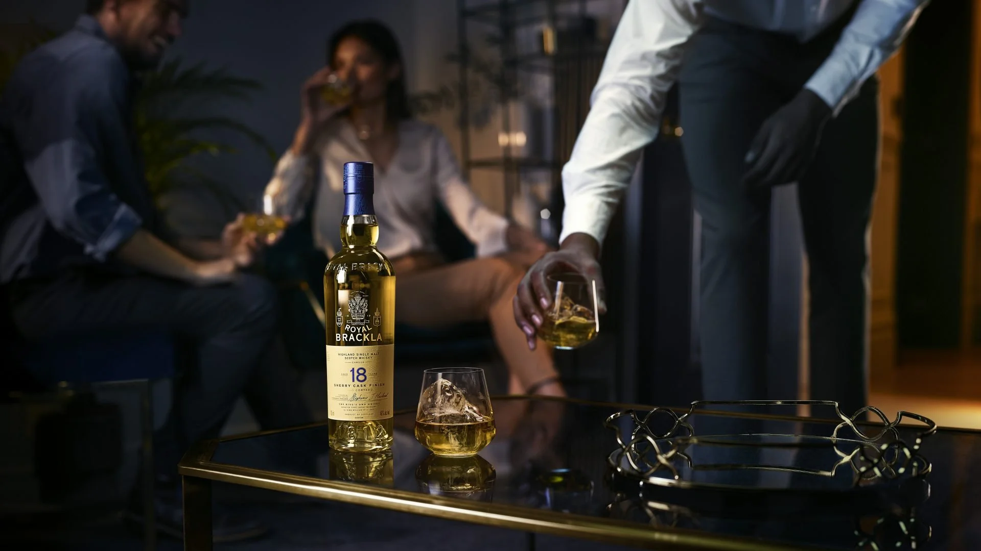 A bottle of Royal Brackla whiskey and two glasses with ice and whiskey on a black table in a dimly lit room. In the background, three people are socializing and one is pouring whiskey into a glass.