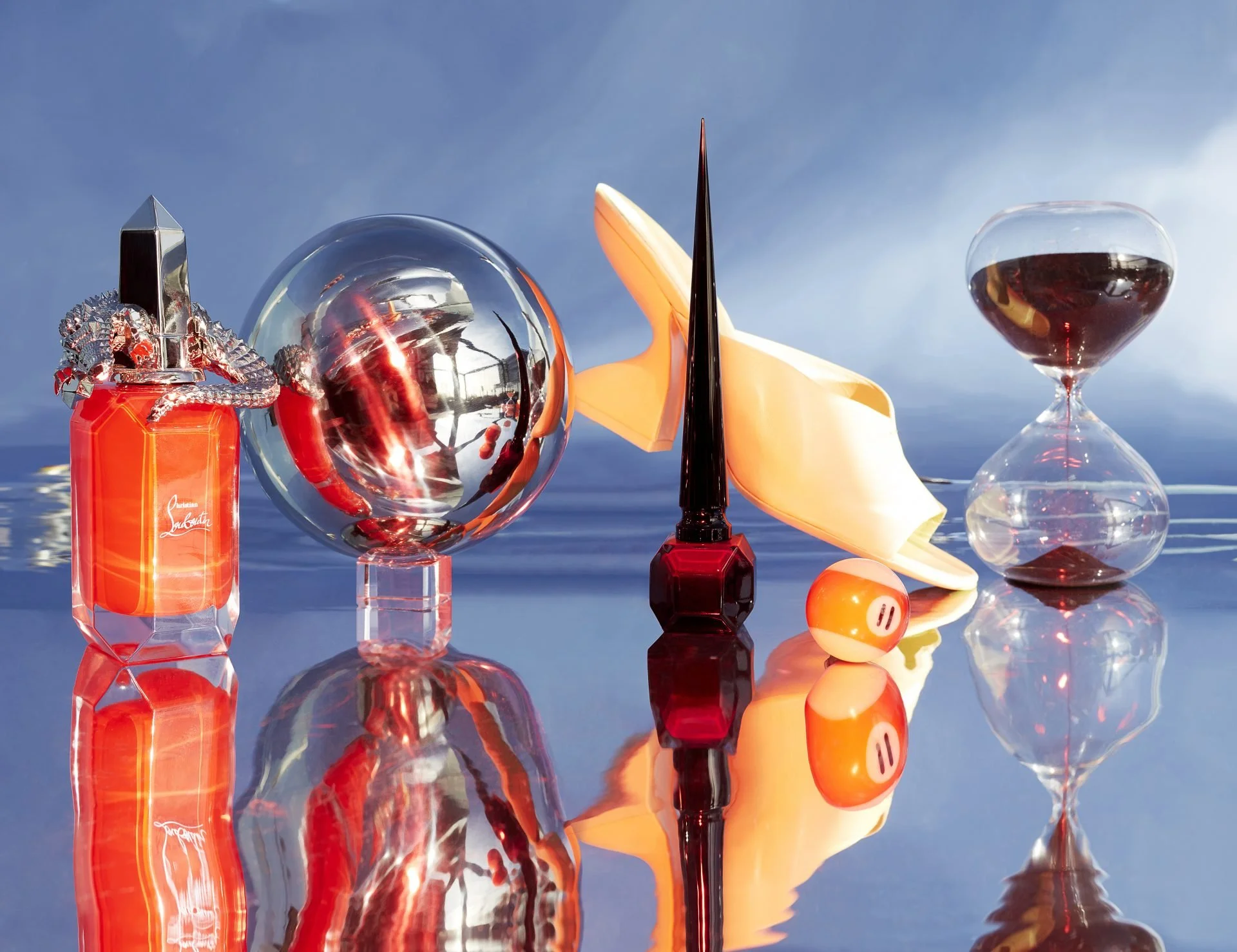Assorted perfumes, a glass sphere, a champagne glass filled with dark liquid, a yellow and orange pool ball, and a dark red nail polish bottle with a pointed applicator, all arranged on a reflective surface outdoors with a blue sky background.