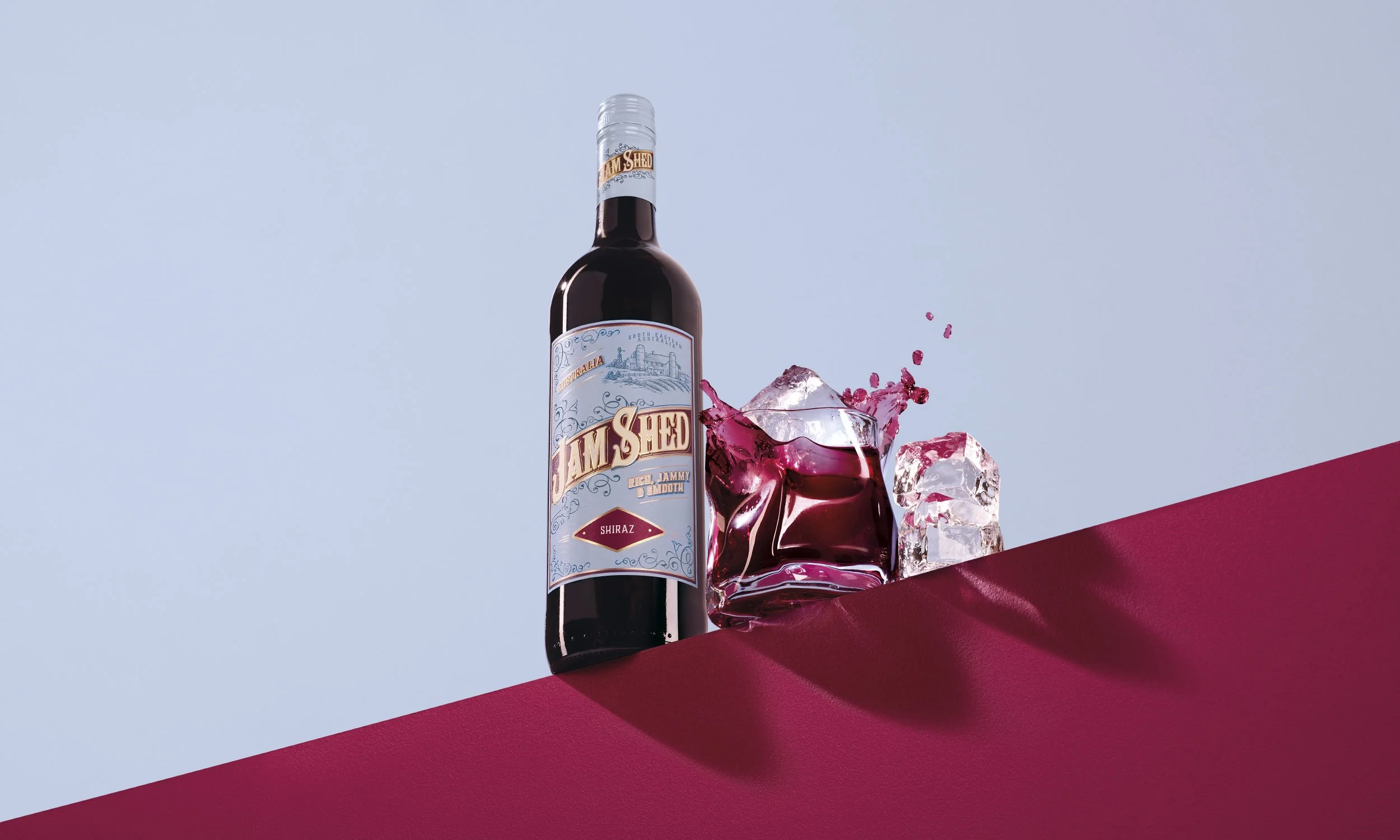 A bottle of Jam Shed Shiraz wine with ice cubes and splashes of red liquid on a red and blue background.