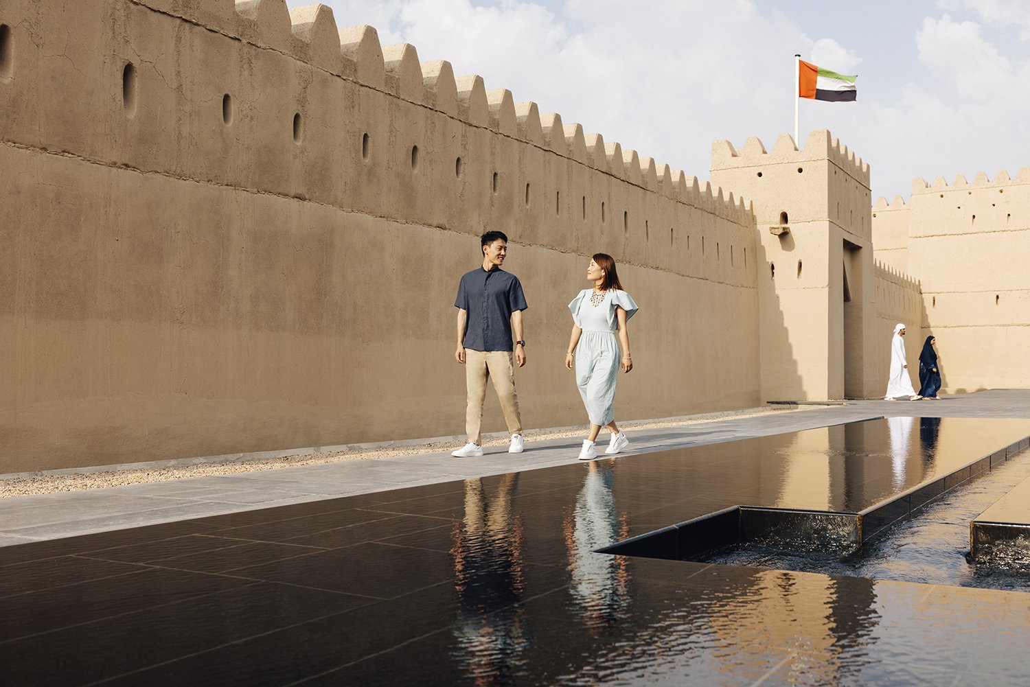 Two tourists walking near a traditional Middle Eastern fort with beige walls, a guard tower, and a flag flying on top. Two other women in traditional attire walk further in the background, with reflections visible in a water feature on the ground.