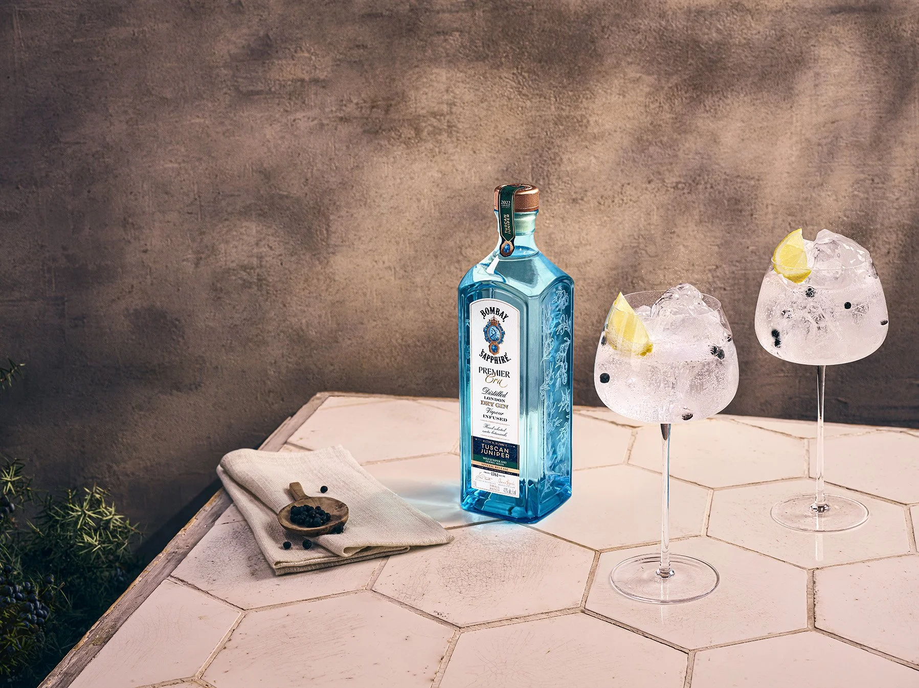 A bottle of Bombay Sapphire gin and two cocktail glasses with ice, lemon wedges, and dark berries on a white hexagonal-tiled surface, with a beige cloth and some berries on a small wooden spoon nearby.
