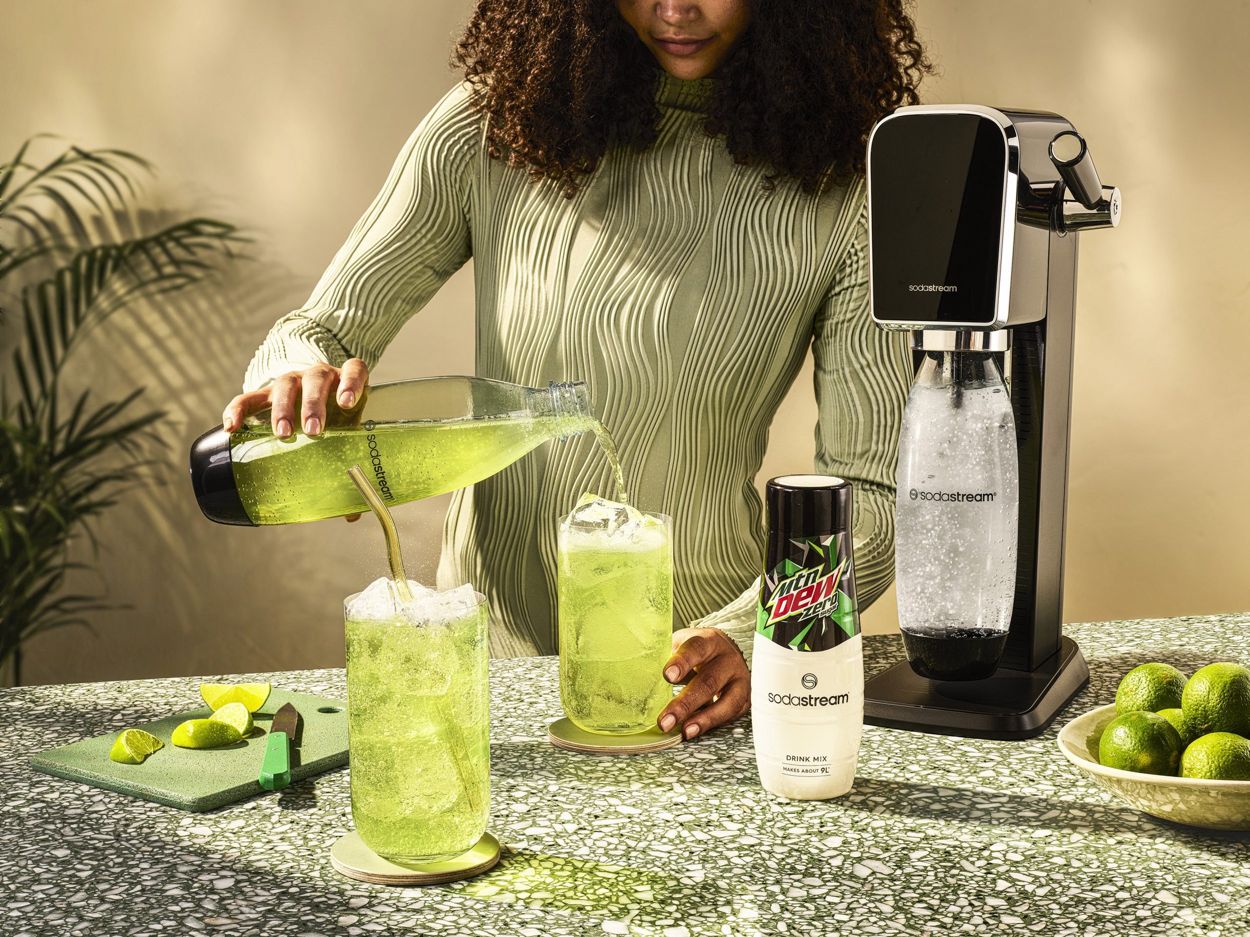 A woman with curly hair preparing a green lime beverage using a SodaStream machine, with limes and lime wedges on the table, and a Mountain Dew Zero Sugar drink in the foreground.