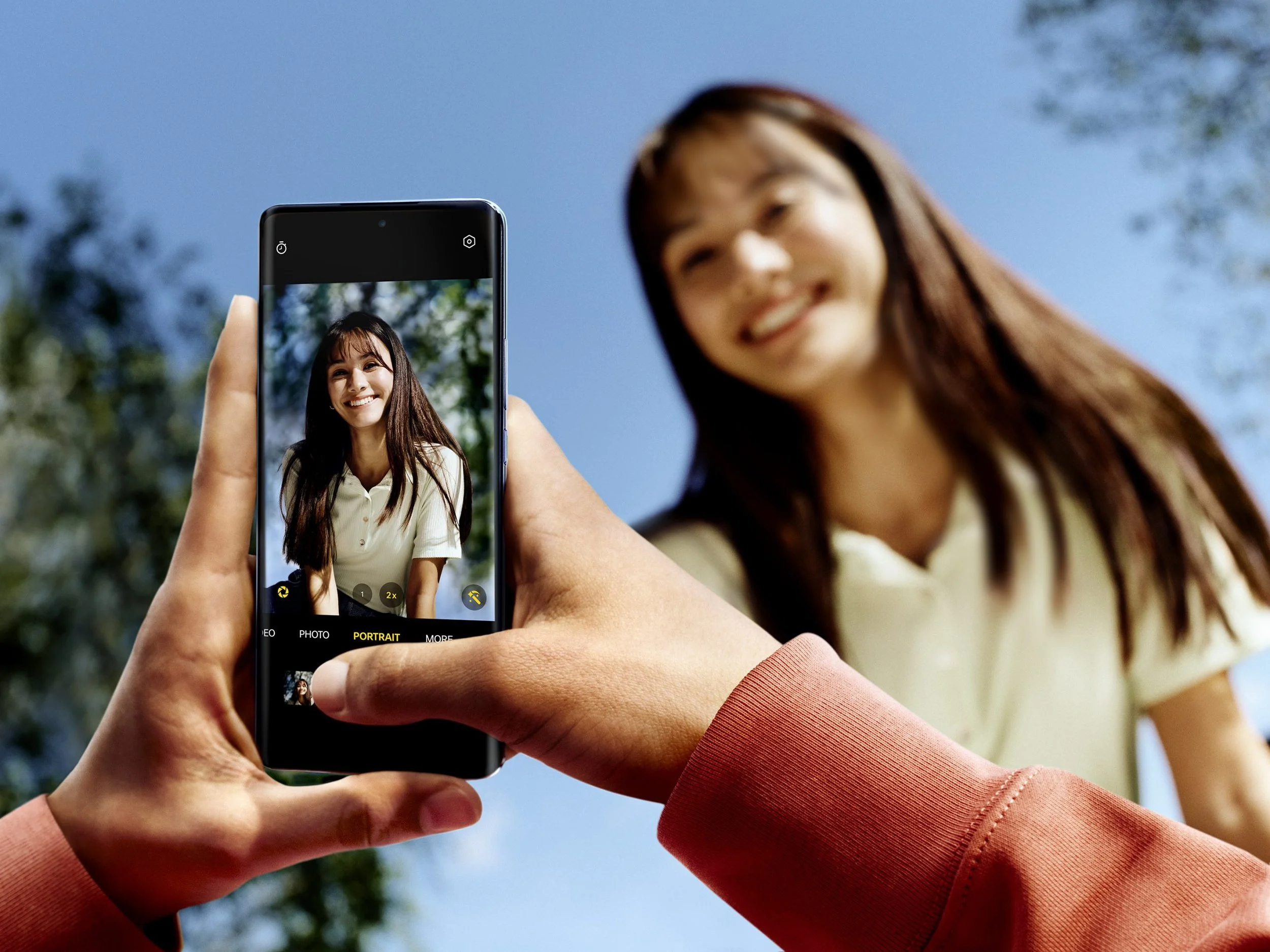 Close-up of a person's hand holding a smartphone, capturing a photo of a woman smiling outdoors with a clear blue sky and trees in the background.