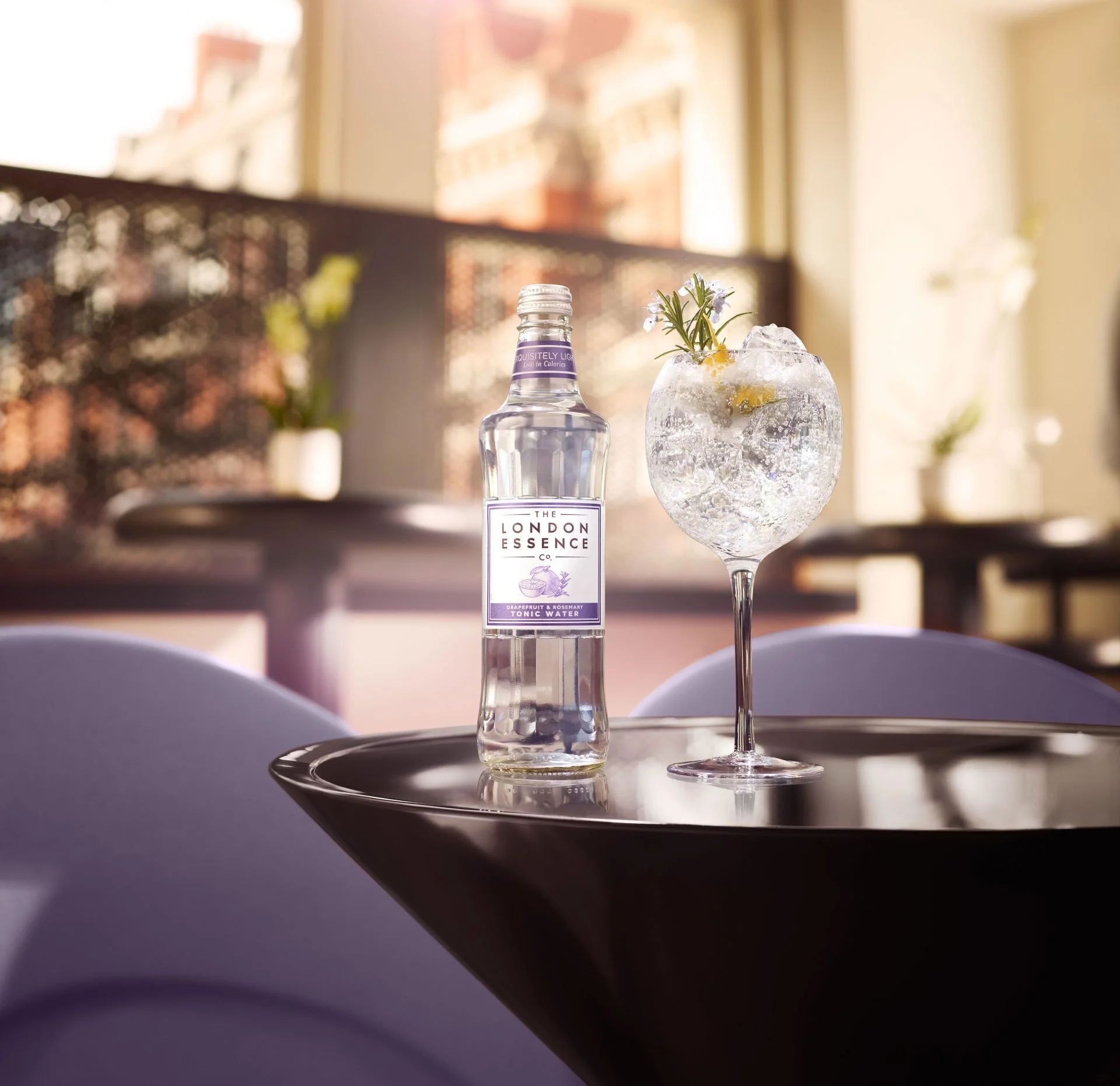 A clear glass bottle labeled "The London Essence Co." with grapefruit and rosemary tonic water, a glass filled with ice, a lemon slice, rosemary sprig, and edible flowers, set on a reflective black tray in a brightly lit room with windows in the back