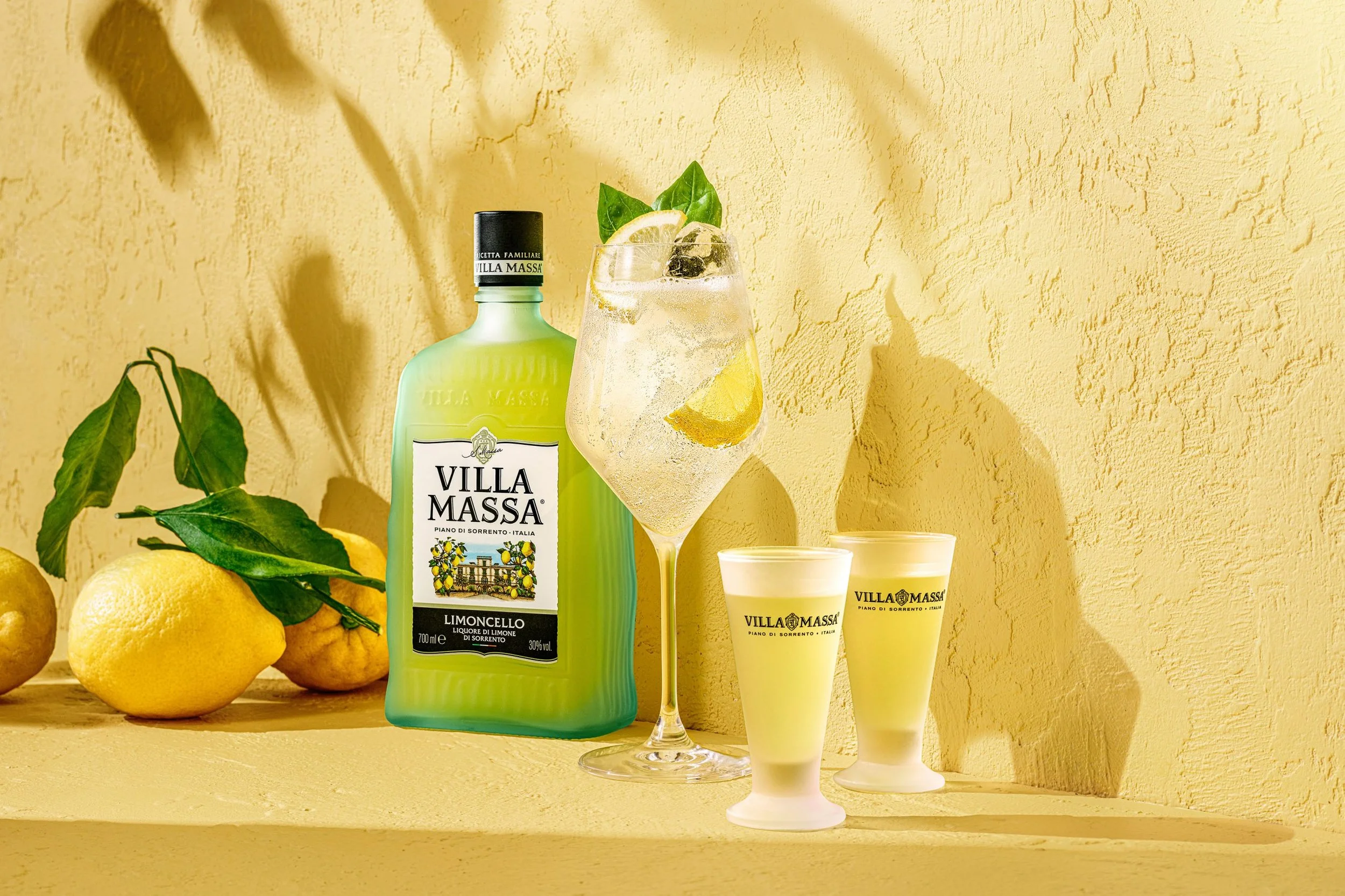 A bottle of Villa Massa Limoncello with a glass of lemon-flavored cocktail garnish, surrounded by lemons and green leaves on a yellow textured background.