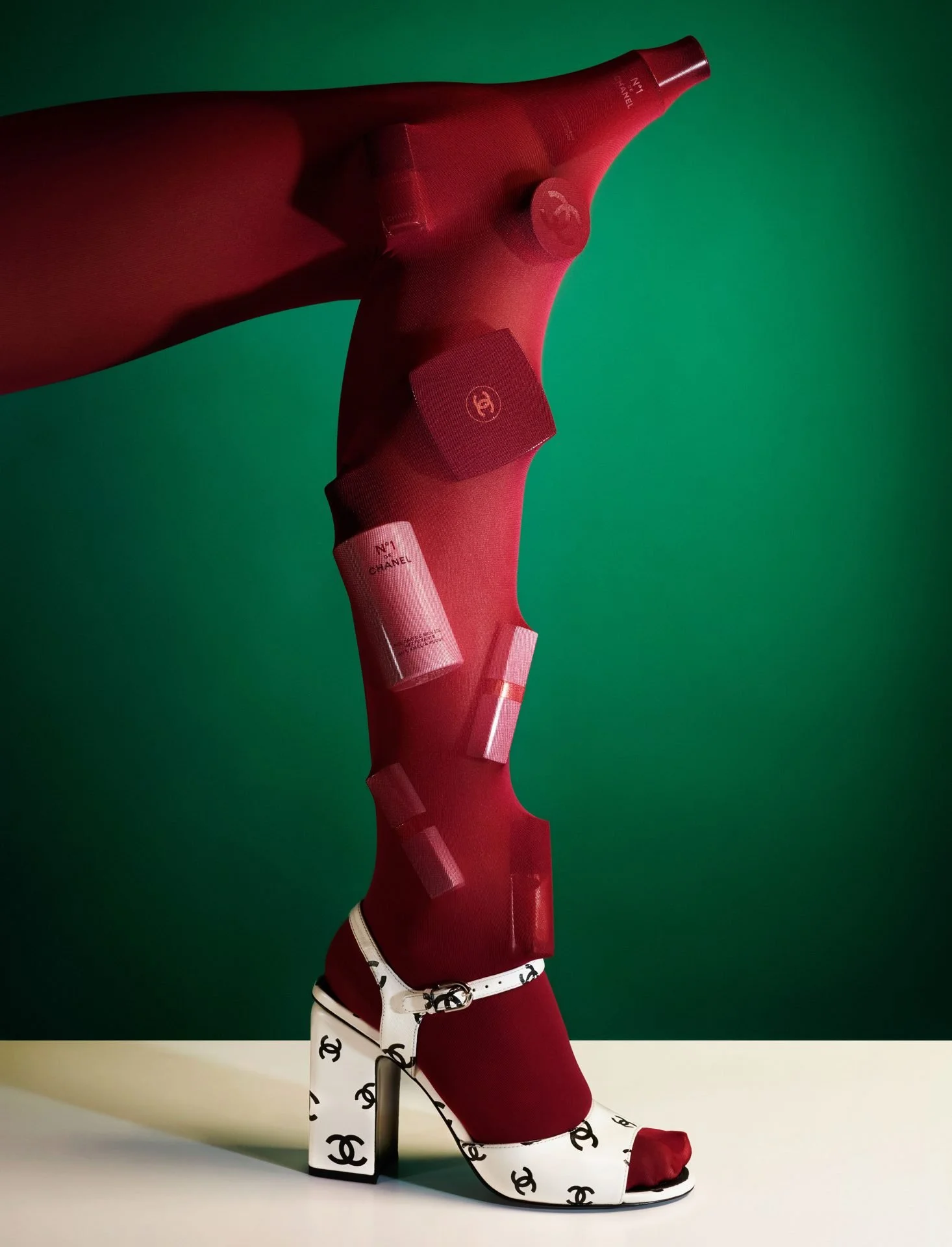 A mannequin leg dressed in designer fashion, including a white high-heeled shoe with black 'Chanel' logos, a red stocking, and a red leather ankle strap. The background features a vibrant green wall.