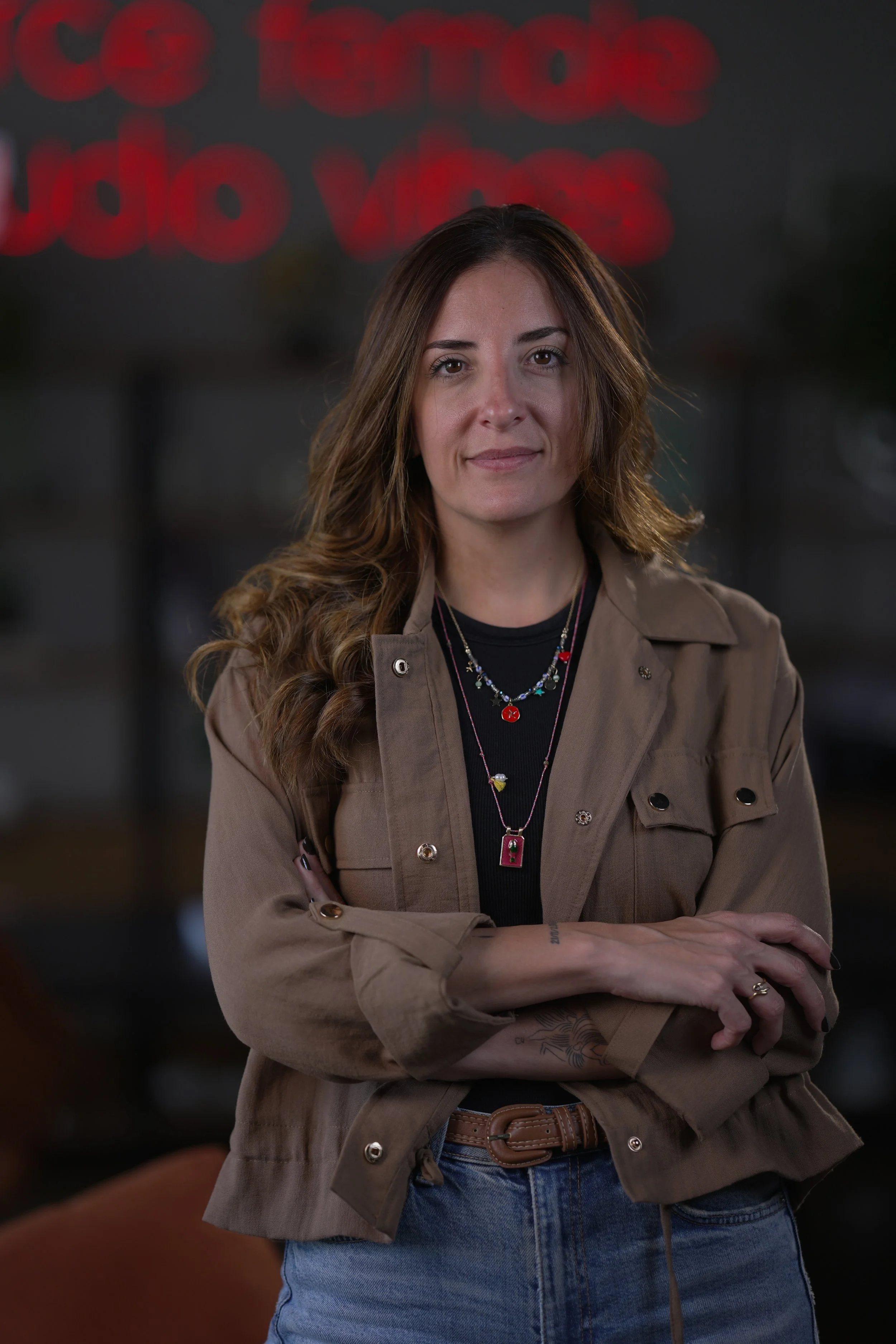 A woman with long wavy brown hair and a tattoo on her forearm, wearing a tan jacket, t-shirt, and multiple necklaces, is crossing her arms in front of a blurred background featuring a neon sign that reads 'coffee'.