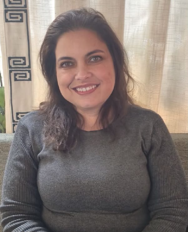 A woman with dark brown hair, smiling, wearing a dark gray sweater, seated indoors with beige curtains behind her.
