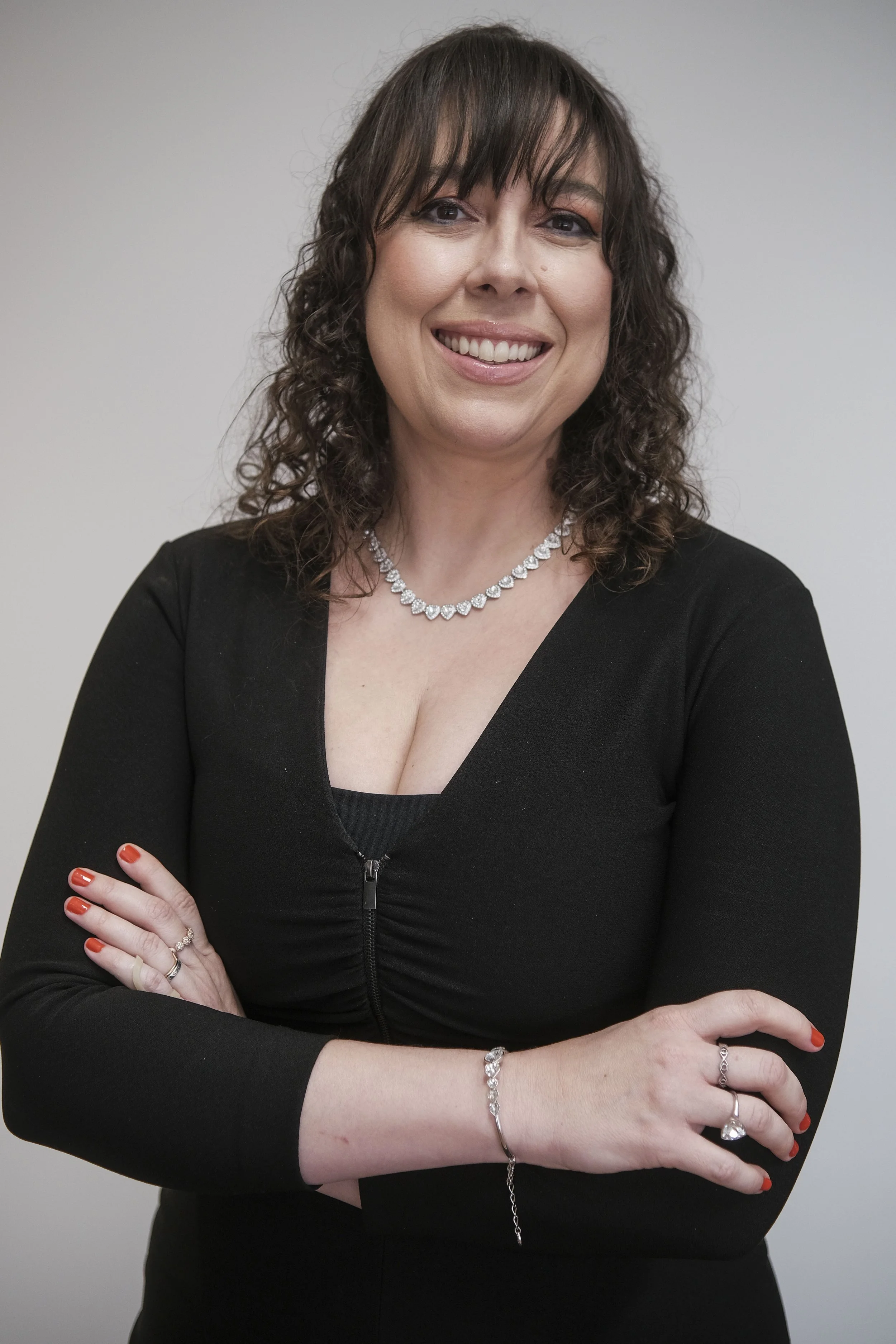 A woman with curly brown hair wearing a black top, a diamond necklace, and matching jewelry, smiling and crossing her arms in front of a plain gray background.