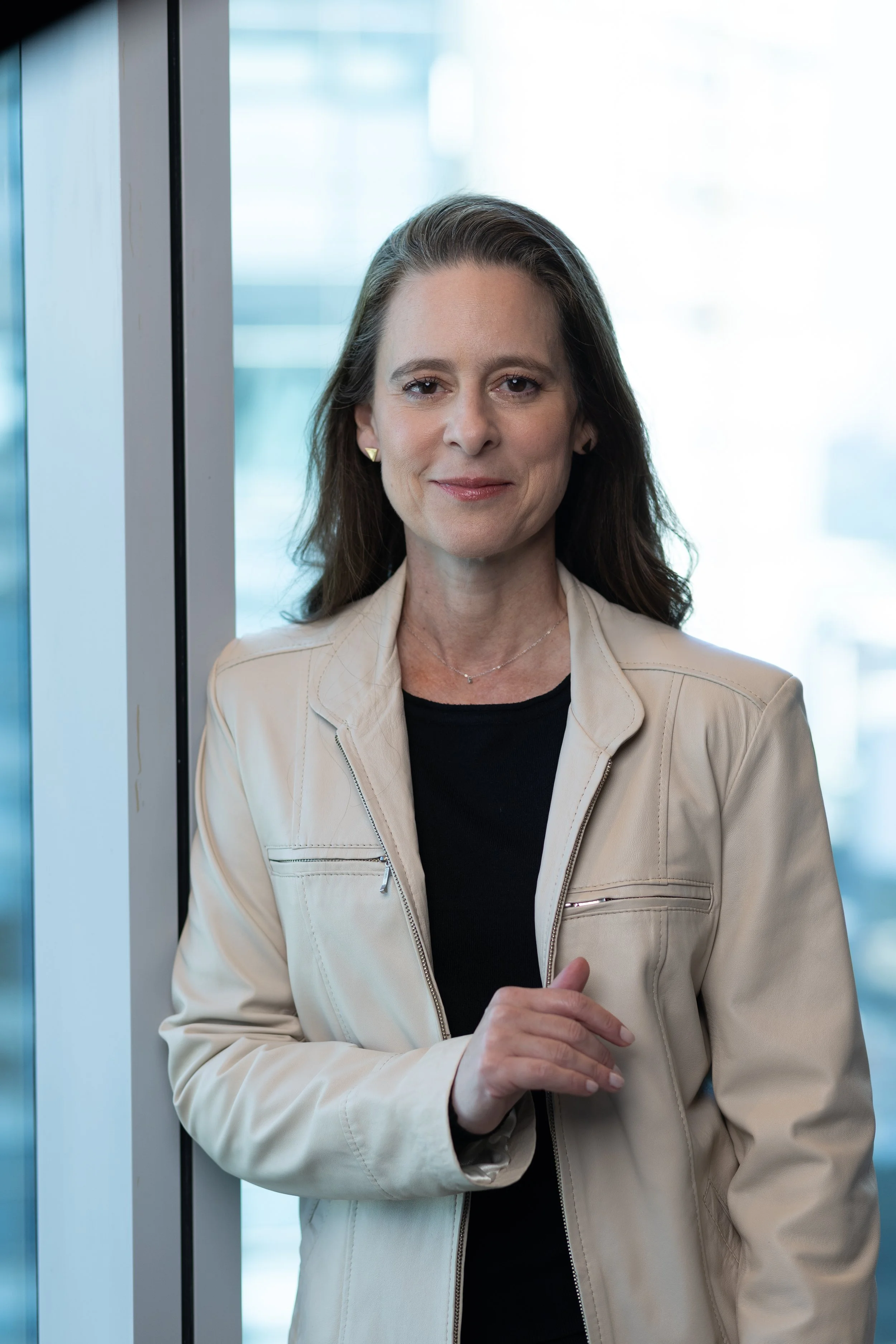 A woman with brown hair, wearing a light beige jacket and black shirt, standing in front of a window with a cityscape background.