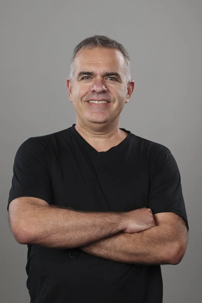 A smiling middle-aged man with short gray hair, wearing a black T-shirt, arms crossed, against a plain gray background.