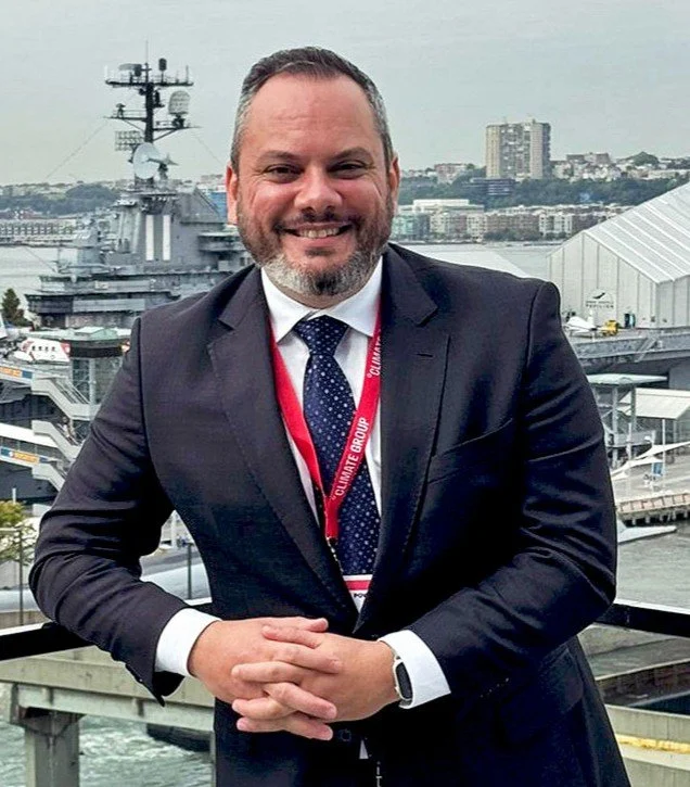 A man in a suit smiling outdoors with a harbor and ships in the background.