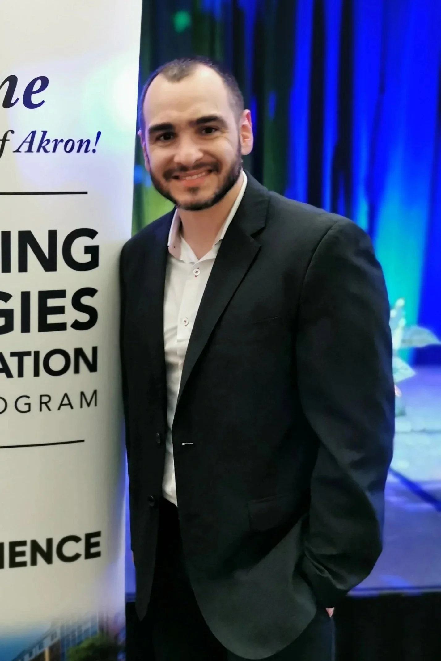 A man with a beard and short hair, dressed in a tuxedo with an open-collared white shirt, standing in front of a sign that reads 'The Future of Akron,' at an indoor event with dark blue curtains in the background.
