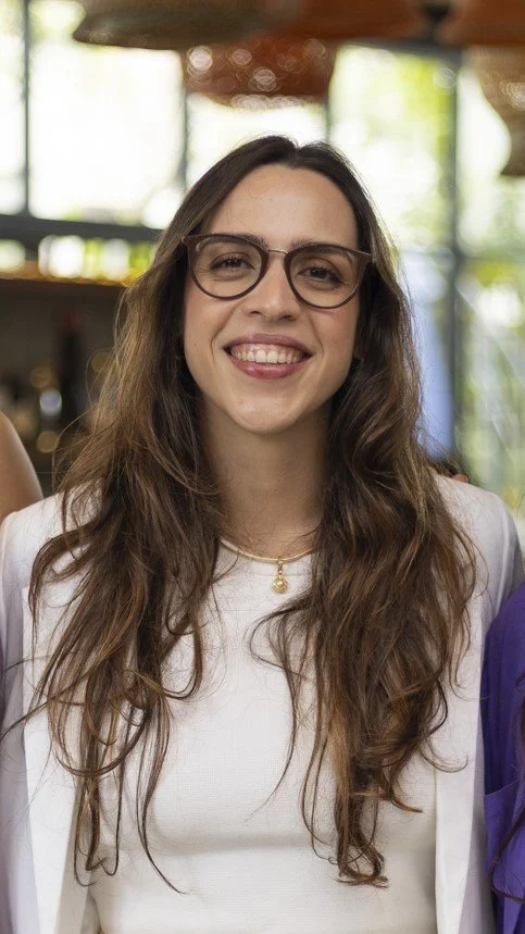 A woman with long wavy brown hair, wearing glasses and a white top, smiling indoors with a blurred background of greenery and wooden furniture.