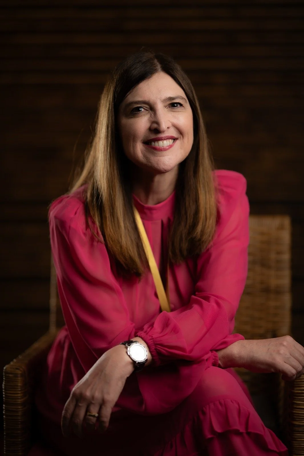 Portrait of a woman with long brown hair wearing a pink blouse sitting on a wicker chair