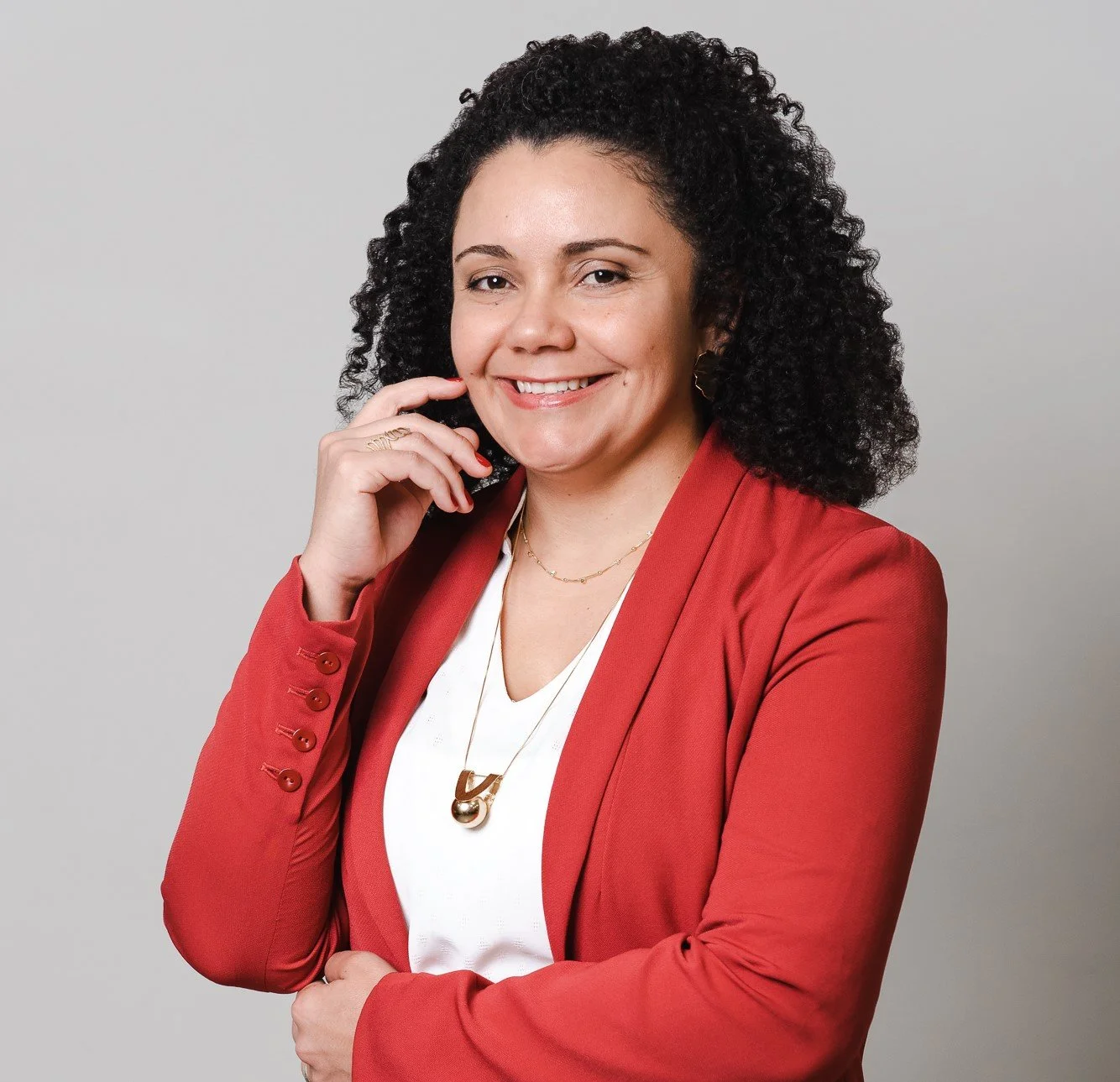 A woman with curly dark hair smiling, wearing a red blazer and white top, with gold jewelry, posing with hand near her face against a plain light gray background.
