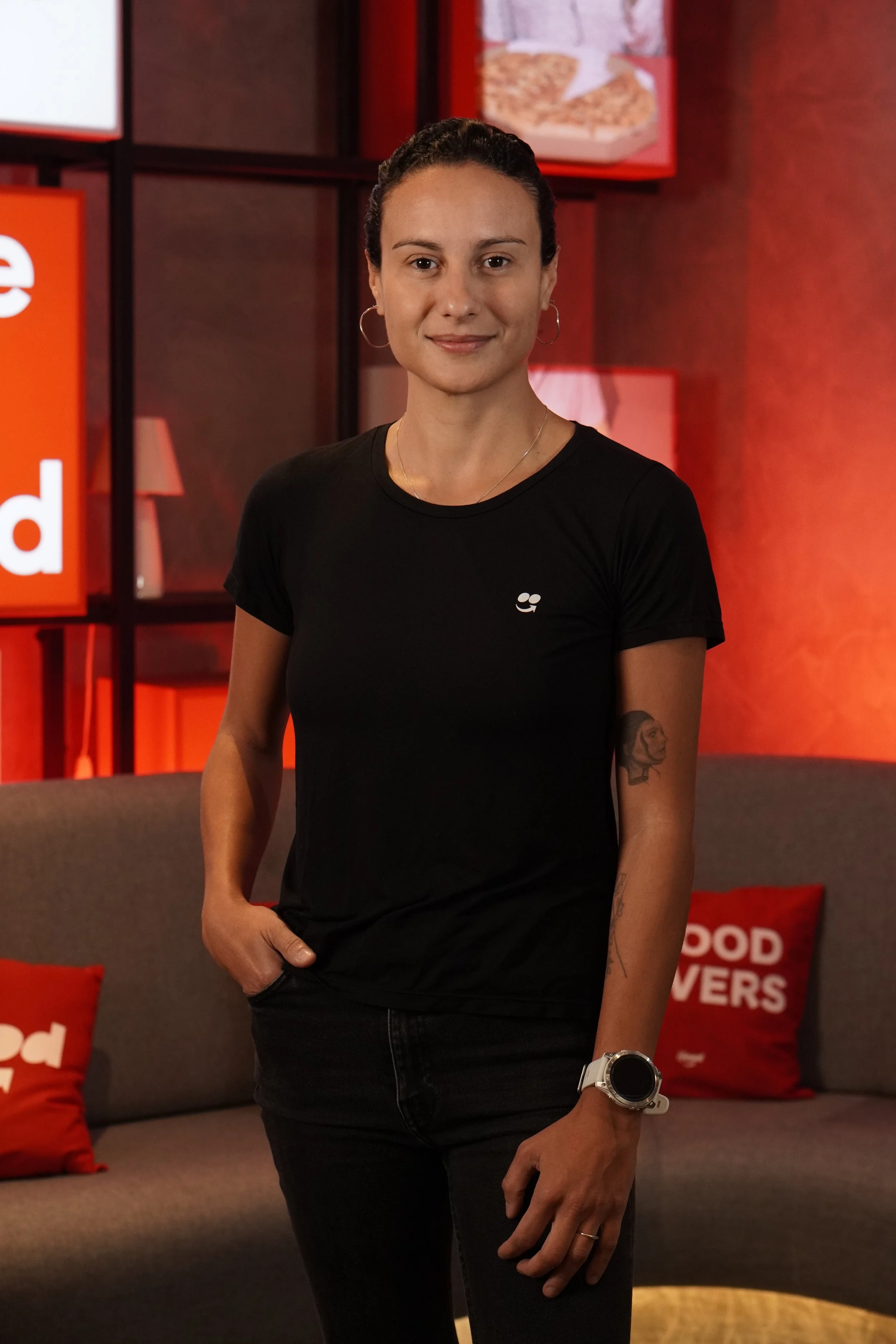 A woman with dark hair pulled back, wearing a black T-shirt with a small logo, hoop earrings, and a watch, standing in a room with red and black decor, including red pillows and a sign with partially visible white text.