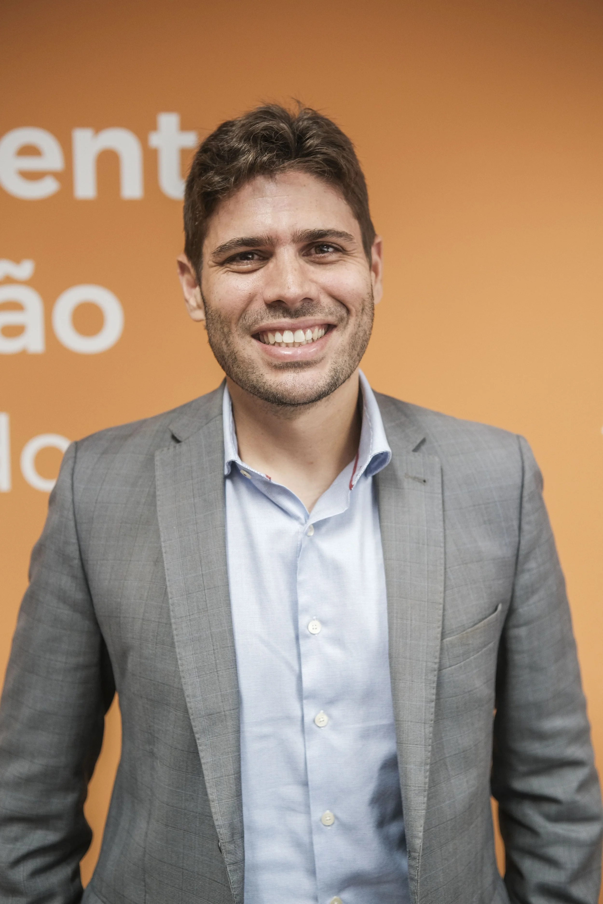 A smiling man wearing a light blue dress shirt and a gray checkered blazer, standing in front of an orange background with partial white text.