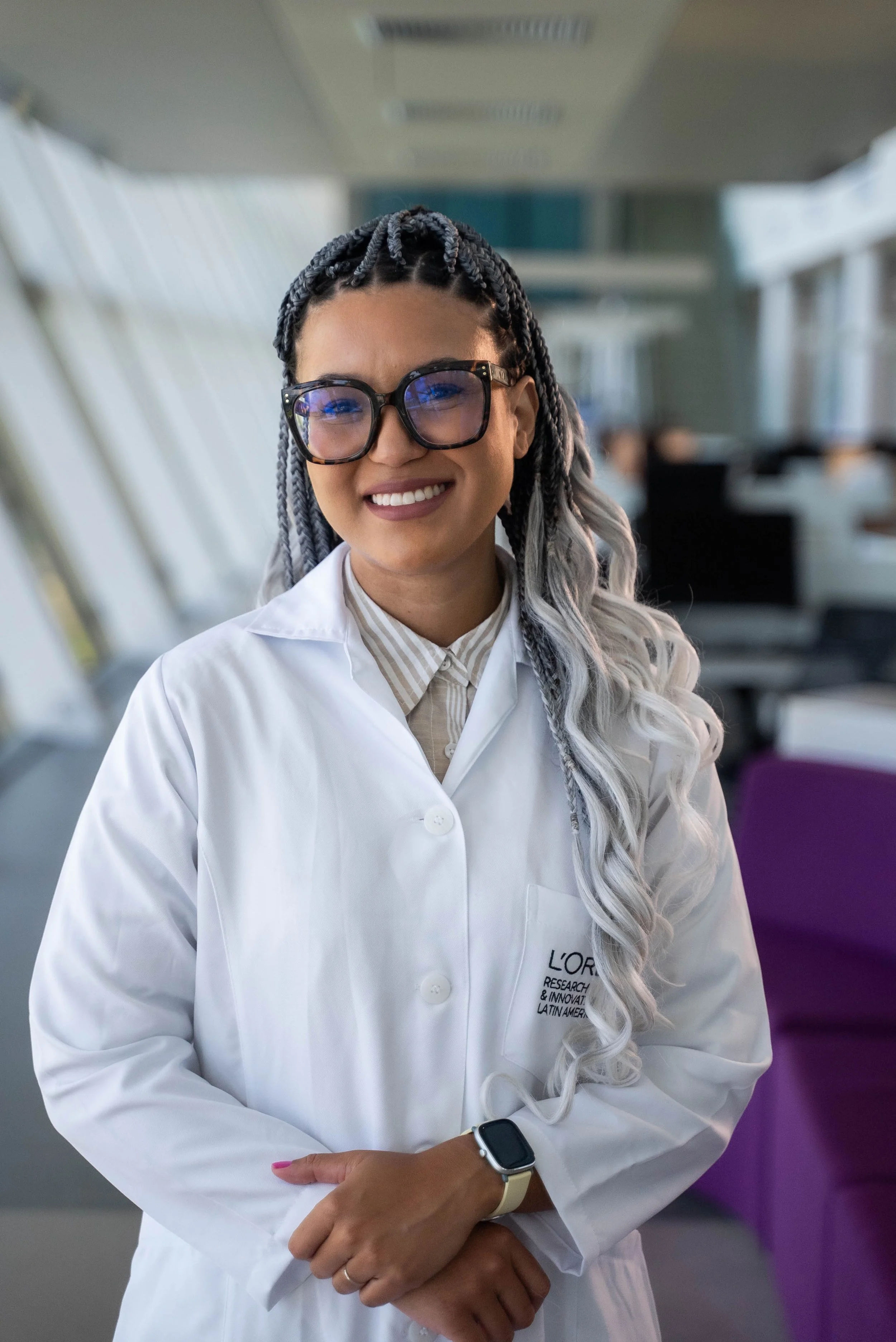 A woman with braided gray hair, glasses, and a white lab coat standing in a modern, brightly lit building with large windows and purple chairs in the background.