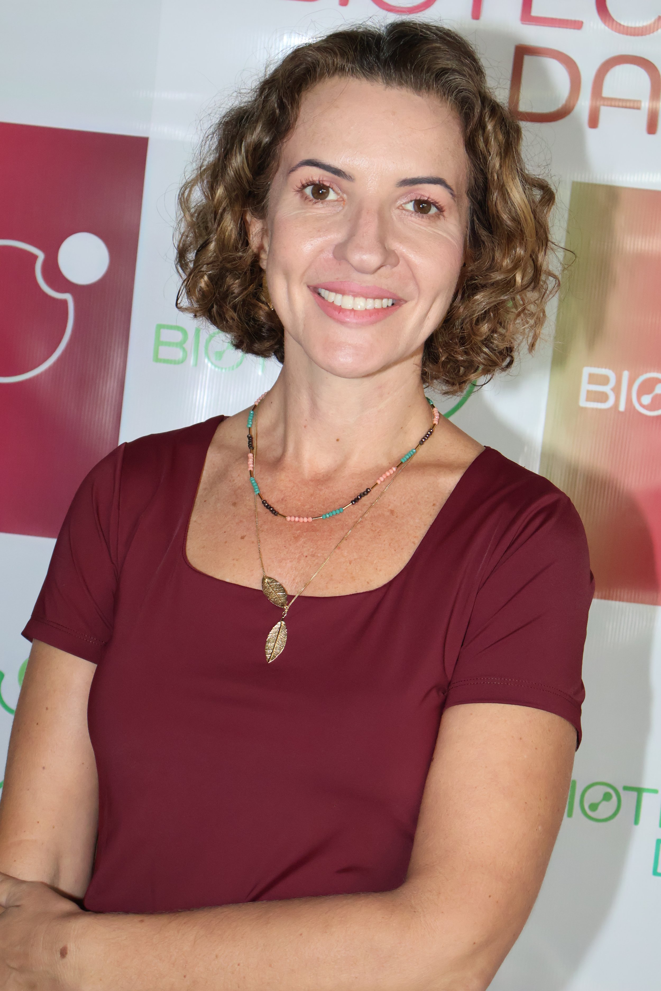 A woman with curly brown hair and brown eyes, smiling, wearing a maroon dress and layered necklaces, standing in front of a backdrop with logos and text.