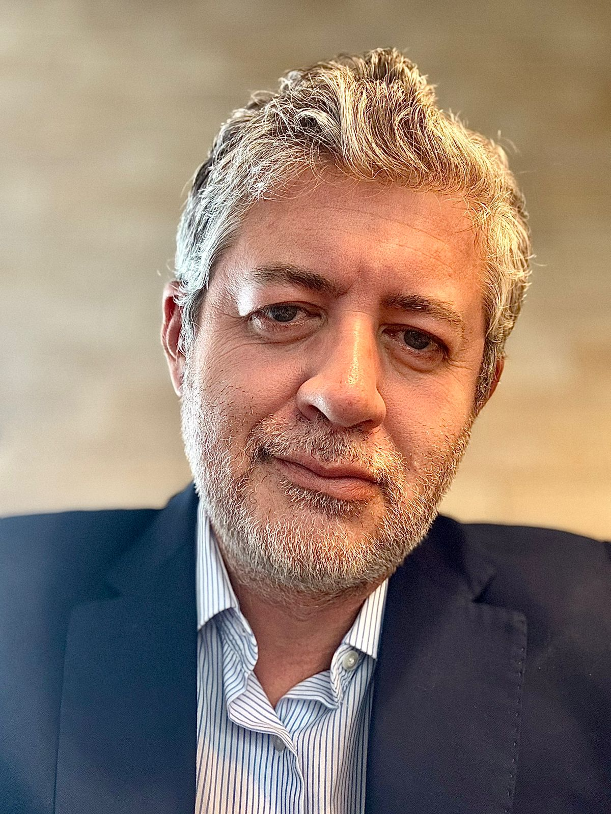 A middle-aged man with grey hair and a beard, wearing a dark suit jacket and a striped shirt, looking at the camera with a neutral expression.